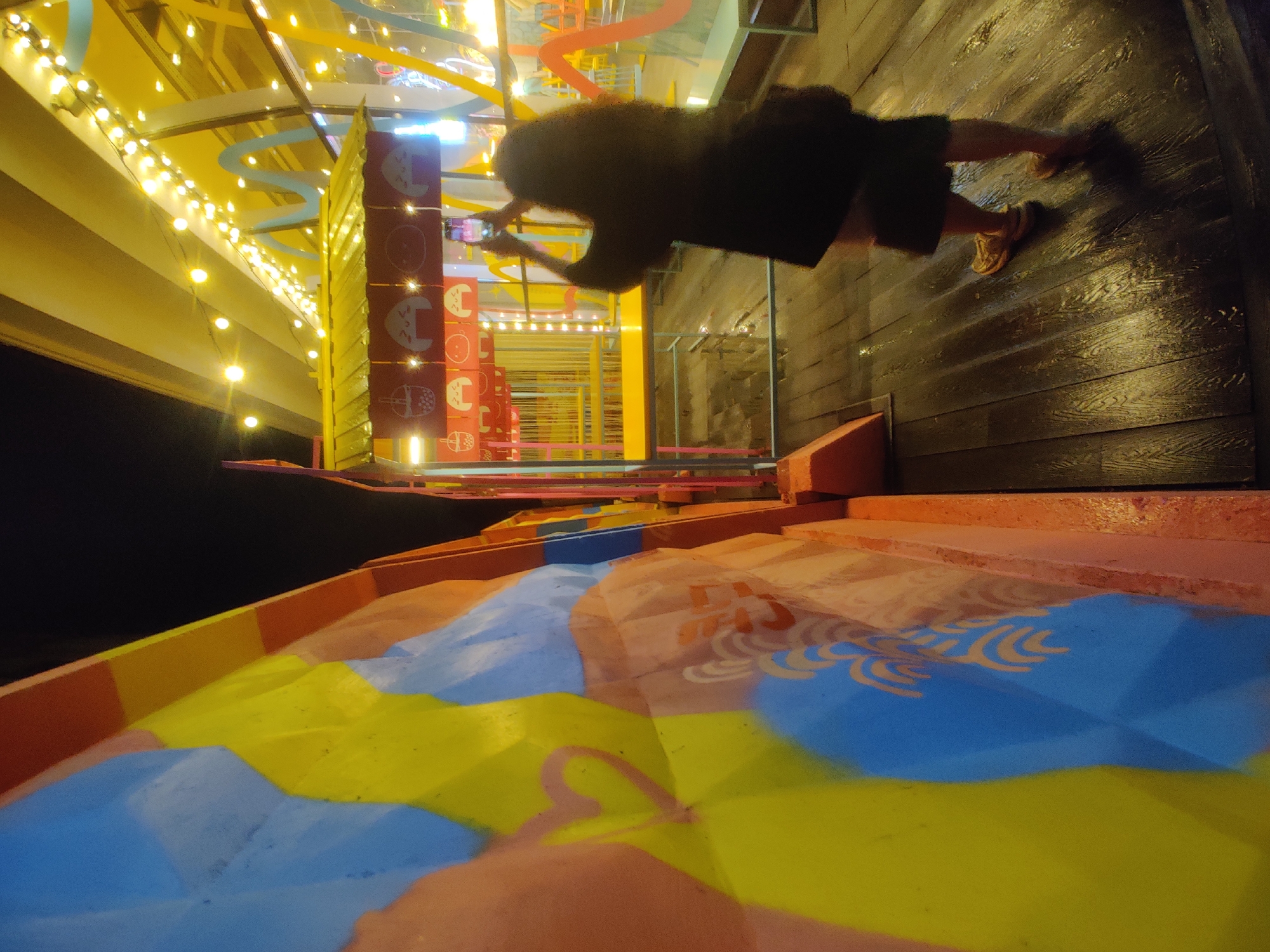 The image depicts a person standing on a wet wooden deck, holding up a smartphone to take a picture. The person is wearing a dark jacket and shorts, and appears to be photographing a brightly colored structure with yellow and red accents, adorned with string lights. To the left of the frame is a wall with a vibrant mural featuring large abstract shapes in yellow, blue, and red, along with a pink heart symbol. The background suggests an outdoor market or food stall area, with more colorful structures and lighting visible. The overall atmosphere is lively and somewhat festive, despite the wet ground, which indicates recent rain or a damp environment. There is no identifiable information about the specific location in Yekaterinburg, Russia, in the image.