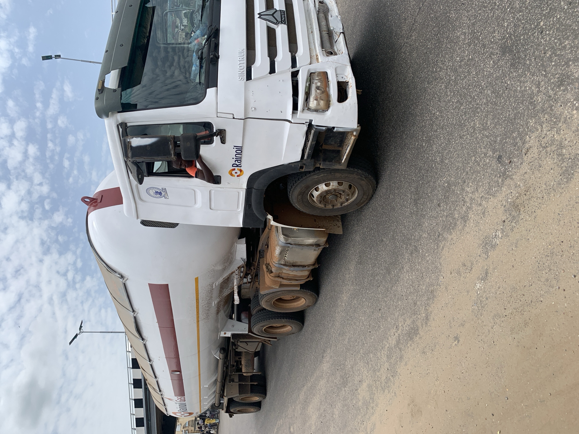 A white SinoTruk commercial vehicle is on a road in Lokoja, Nigeria. The vehicle is a tanker truck and is marked with the "Rainoil" logo. A person wearing an orange shirt can be seen inside the truck. The road is paved, and there is a light post with two lights visible in the upper left portion of the picture. Buildings are visible in the background. The sky is partly cloudy.