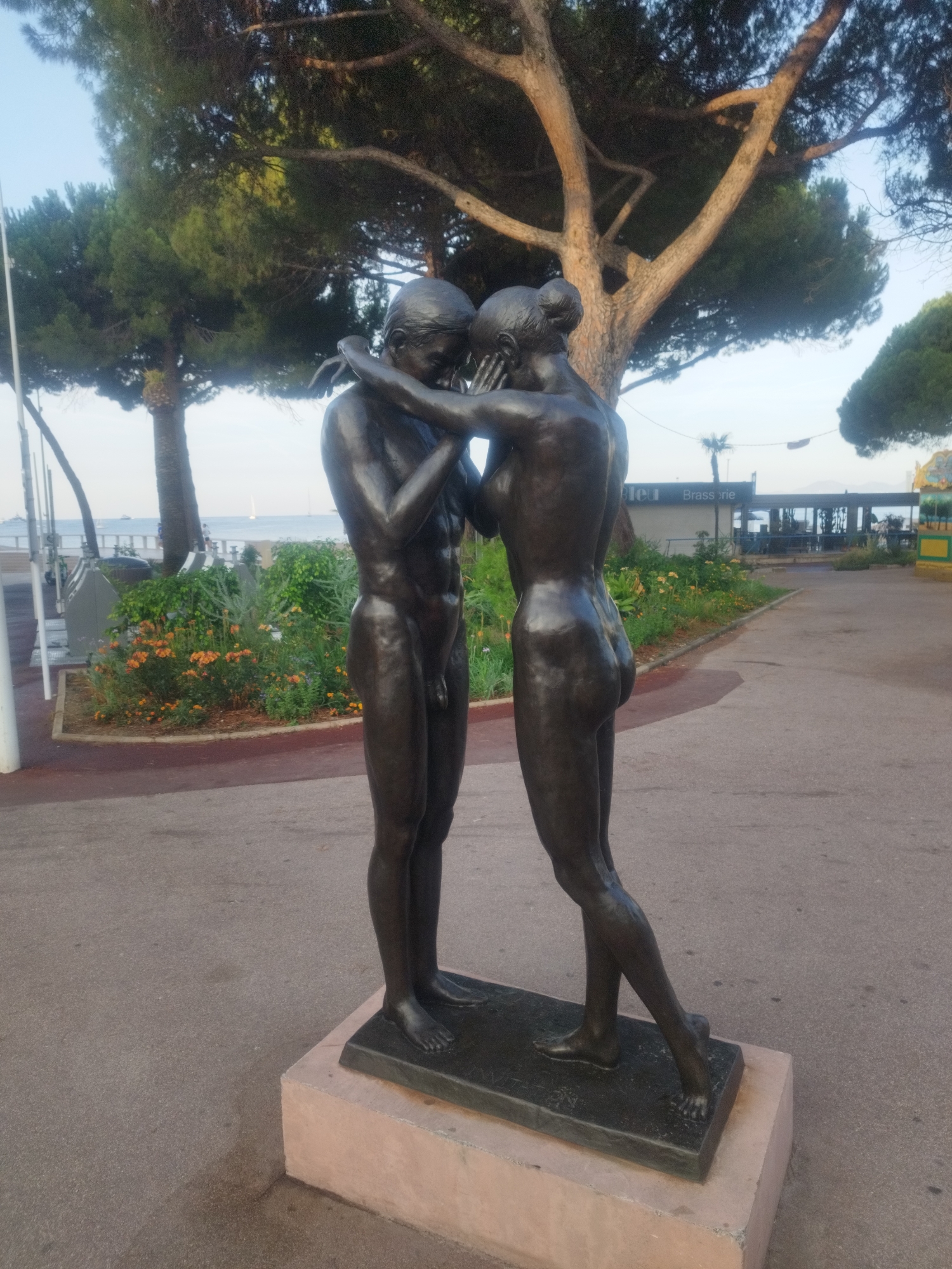 A bronze sculpture depicting two nude figures in an embrace stands on a concrete promenade. The figures are positioned on a rectangular plinth. The male figure is to the left, facing the viewer with his arms around the female figure. The female figure is to the right, facing away from the viewer, with her head turned towards the male. Both figures have a polished finish.
Behind the sculpture, a pathway leads towards buildings and trees. In the background, there is a body of water with sailboats, suggesting a coastal location. A building with the text "Brasserie" is visible. The promenade is paved with asphalt, and there are flower beds with orange and yellow flowers to the left of the sculpture. Tall trees, likely pine trees, with their branches spreading overhead, frame the scene. The sky is visible in the upper part of the image, with hints of blue and overcast clouds. This sculpture is known as "The Kiss" and is located on the Promenade de l'Amiral de Grasse in Antibes, France.