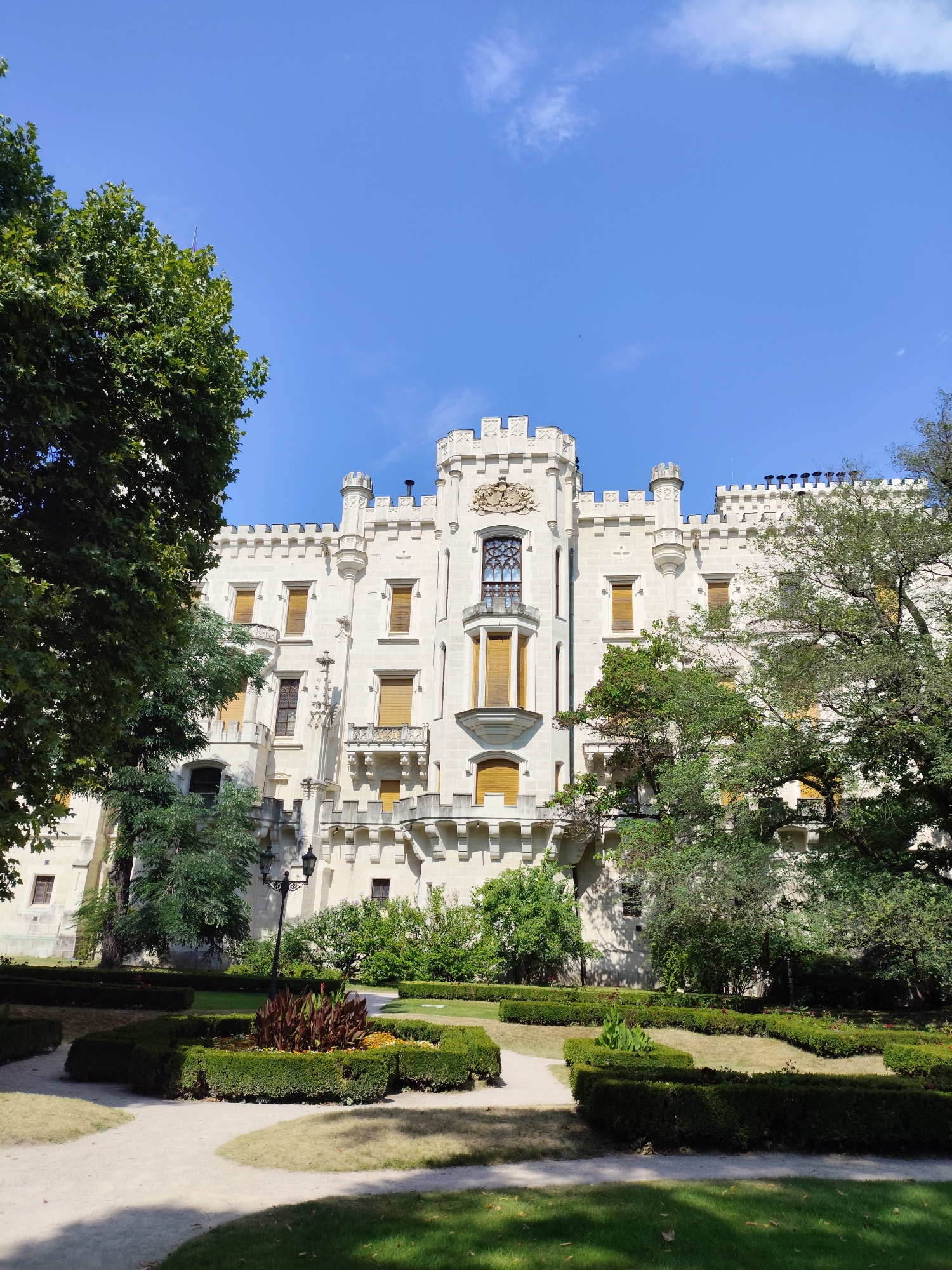 This is a medium-shot, eye-level, outdoor, daytime photograph of Hluboká Castle, located in Hluboká nad Vltavou, Czechia. The castle is a prominent white structure with crenellated battlements and towers, characteristic of Gothic Revival architecture. The building has numerous windows, many with closed wooden shutters. Lush green trees frame the castle on both sides, and a meticulously maintained formal garden is in the foreground. The garden features manicured hedges, curved pathways, and a central flower bed with tall, dark red plants. The sky above is a clear, bright blue with a few wispy white clouds. There are no people or animals visible in the image. The scene conveys a sense of grandeur and tranquility.