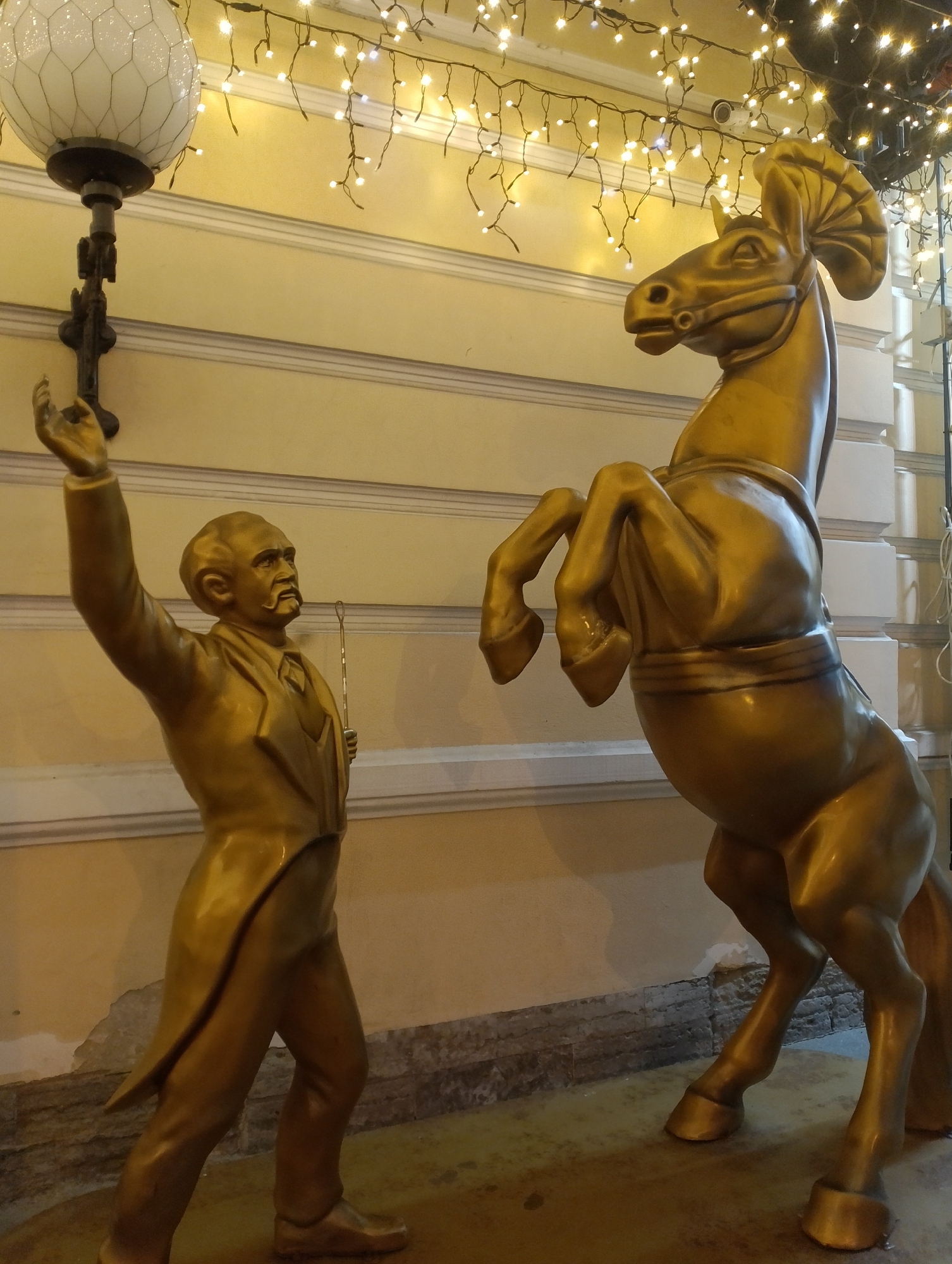 The image depicts a scene in front of a building in Saint Petersburg, Russia, likely during the evening or at night due to the artificial lighting. The primary subjects are two golden statues: a man and a horse.
The man, on the left, is depicted in a suit, with one arm raised as if gesturing or reaching for something. He is holding a thin rod in his other hand. His facial features are detailed, showing a mustache and a stern expression. He appears to be standing on a textured surface, possibly a sidewalk or pavement. Above and to his left is a decorative lamp with a patterned glass shade.
To the right of the man is a statue of a rearing horse, also golden. The horse is positioned dynamically, with its front legs raised off the ground. It is adorned with a bridle and a decorative element on its head, resembling a fan or crest. The horse's body is robust and detailed, emphasizing its muscular form.
The background consists of a building facade with a light yellow color and architectural details like molding. A string of white fairy lights is draped above the statues, illuminating the scene and creating a festive atmosphere. Some of the lights appear to be focused on the building's exterior. The overall impression is that of an outdoor decoration or public art installation.