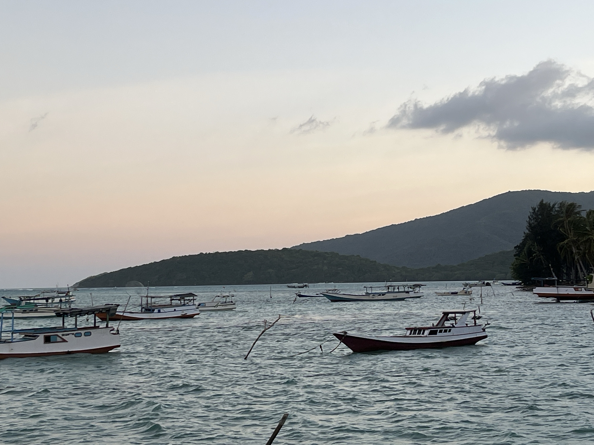 The image shows a harbor in Legonipah, Indonesia, at dusk. Several boats are anchored in the water. The sky is a pale orange and pink, and there are a few clouds. A tree-covered mountain range is visible in the background. The water is choppy, and there are some wooden poles sticking out of the water.