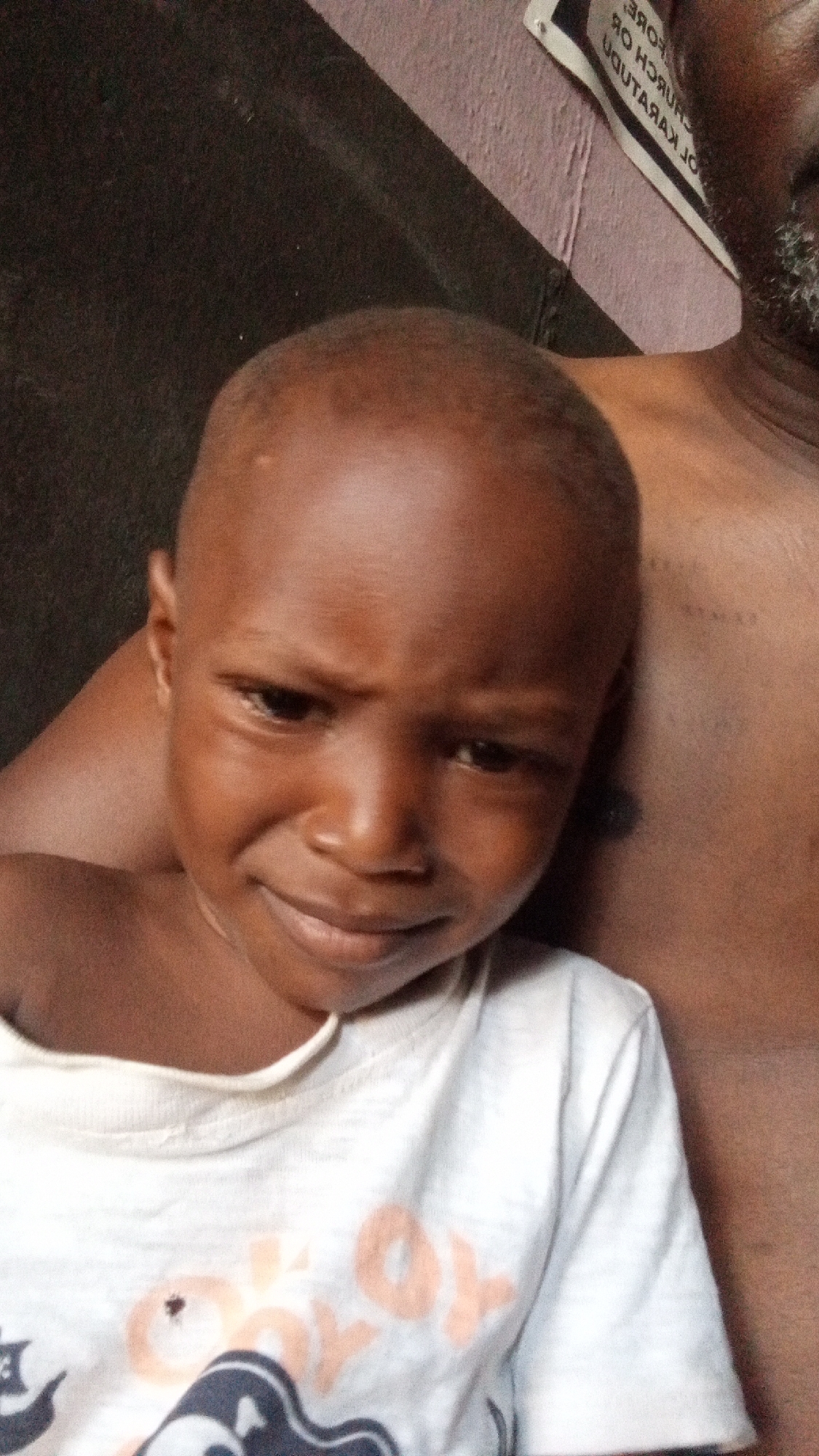 The image is a close-up portrait featuring a young child, likely a boy, with dark skin and a closely shaven head, looking directly at the camera. The child's expression is somewhat serious or thoughtful, with a slight furrow in the brow and a subtle downturn at the corners of the mouth.
The child is wearing a white t-shirt that has faded light orange and dark blue graphic prints on it, including what appears to be the letters "OX" in orange. The child is positioned in front of an adult, whose bare shoulder and chest are visible on the right side of the frame. The adult appears to have some grey stubble or a beard visible in the upper right corner, partially obscured.
The background is dark and indistinct on the left side. On the right, behind the adult, there's a wall that appears to be a light purple or pink color. A white sign or piece of paper is attached to this wall, featuring inverted black text. The visible text reads "ЭЯОНƎ", "ЯUНЯUНƆ ꓠO", "UⱯЯAꓘ Oᒋ". When inverted and read from right to left, this text likely translates to "BEFORE CHURCH OF KARATU" or a similar name, consistent with the given location context of Sabon Tasha, Nigeria. The lighting suggests an indoor or shaded outdoor setting.