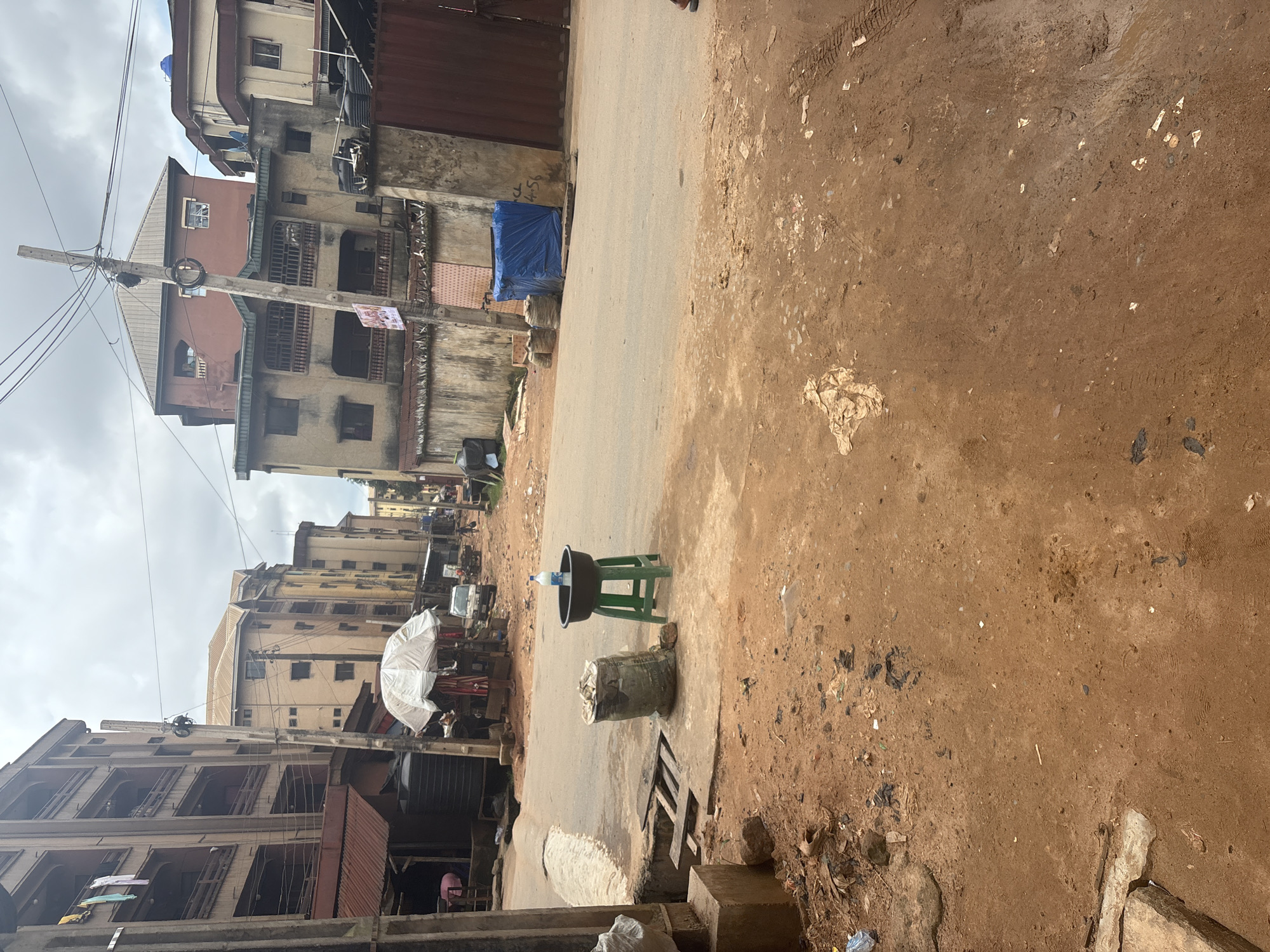 In Onitsha, Nigeria, a street scene unfolds with a mix of urban structures and natural elements. A paved road, marked by tire tracks and scattered debris, cuts through the scene. On the left, a large, dark water tank sits beside a building with multiple balconies, where clothes are hung out to dry. Nearby, a white umbrella covers what appears to be a vendor's stall, with a small white truck parked behind it. Further down the street, other buildings of varying heights and colors line the road.
In the foreground, a large sack, possibly filled with grain or sand, rests on the dusty, reddish-brown earth. Beside it, a green plastic stool supports a dark basin holding a bottle of water, suggesting a makeshift water station or vendor. The ground is uneven, with patches of exposed earth and scattered litter. The sky is overcast, with visible power lines crisscrossing overhead, indicating the presence of electricity infrastructure. The overall impression is one of a busy, lived-in urban environment with signs of commerce and daily life.