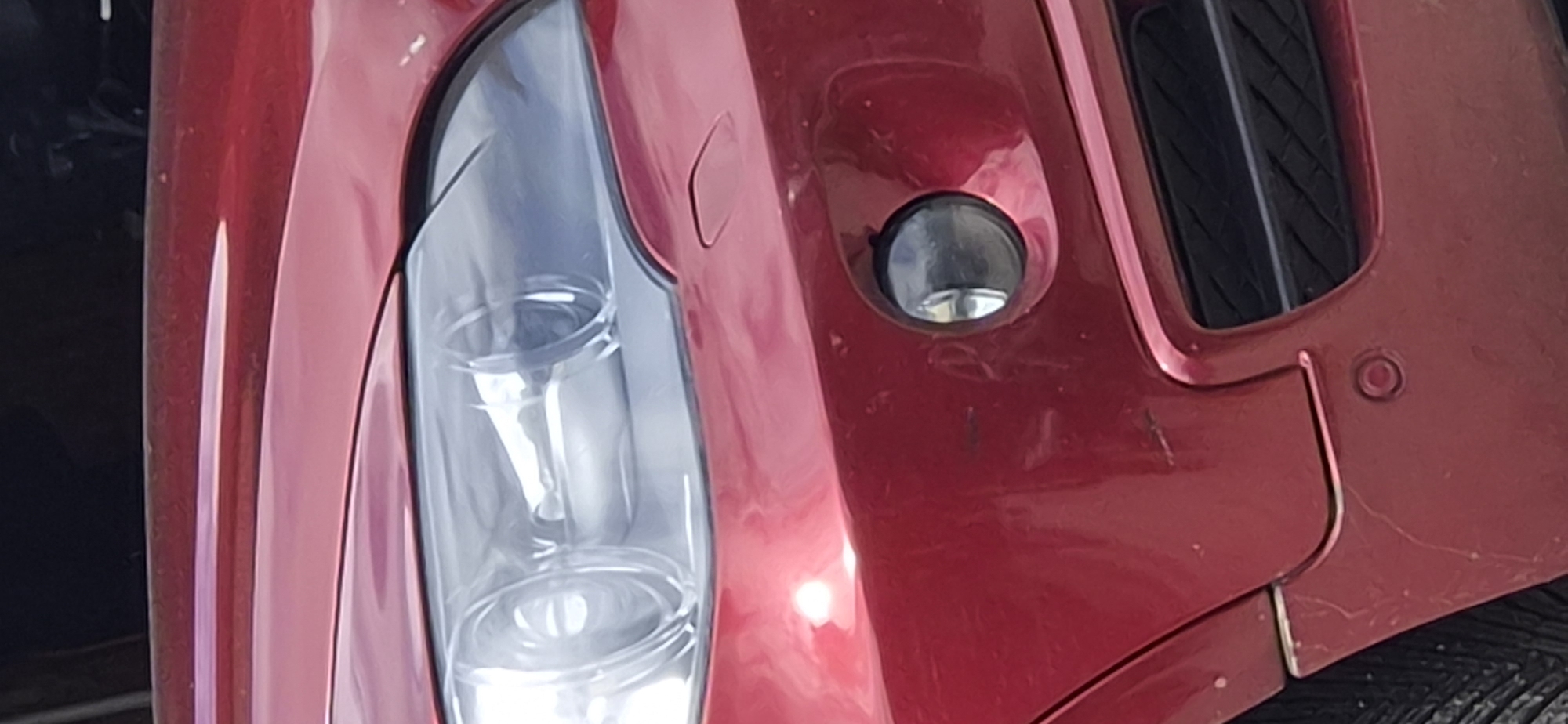 The image shows a close-up of the front passenger side of a red car. The car has a headlight with multiple circular elements and a fog light below the headlight. A black grille is visible in the lower part of the image. A round sensor is mounted on the bumper below the fog light. The car's paint shows some minor scuffs and scratches. The background is dark and out of focus, suggesting an outdoor setting. The scene is not explicitly linked to Radilovo, Bulgaria, but it depicts a common urban or suburban environment where cars are typically found.