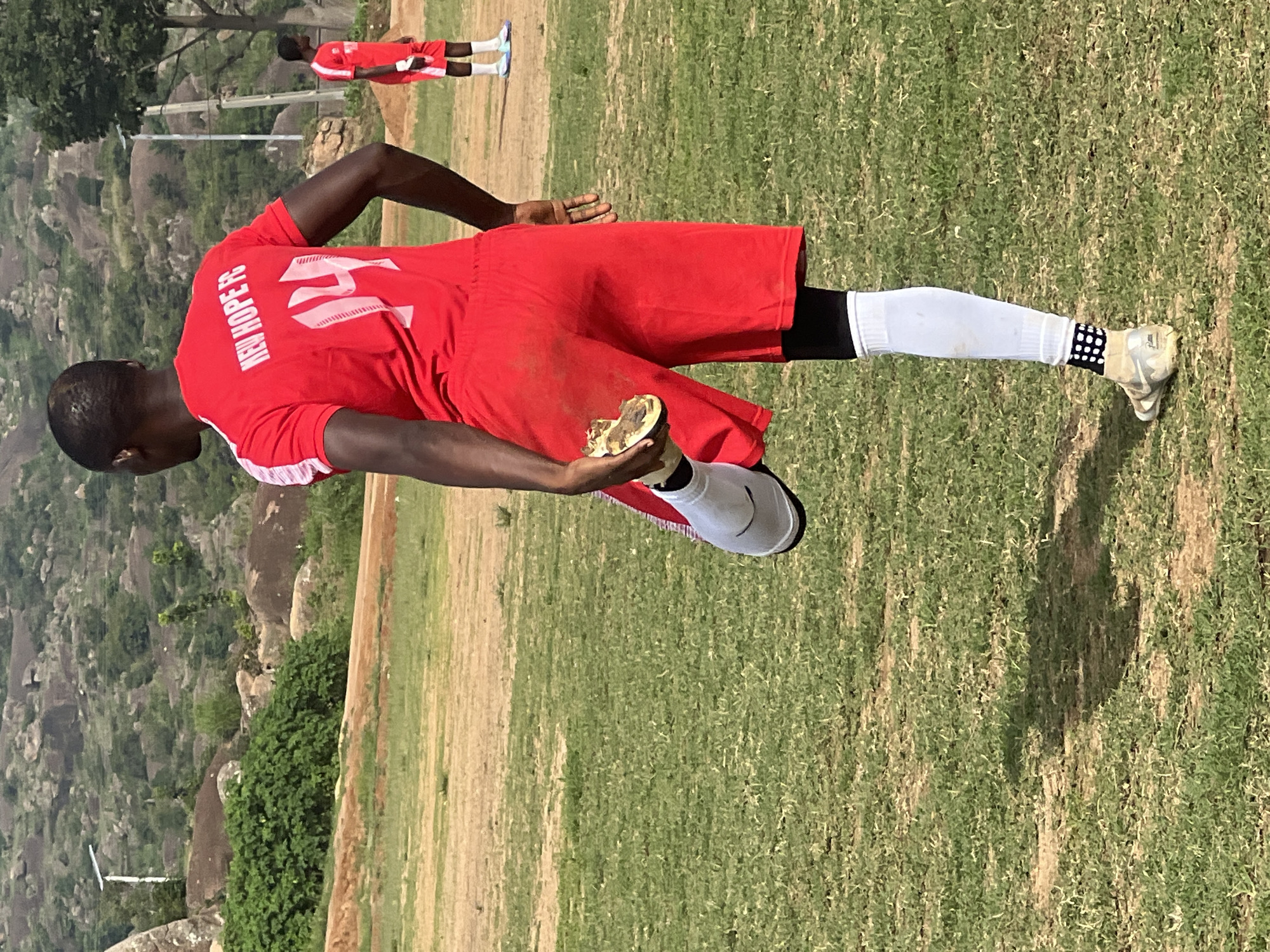 A Black male is seen from behind, wearing a red jersey with "NEW HOPE FC" and the number "14" on the back, and red shorts. He is performing a stretch on a grassy field. His right leg is bent at the knee, with his foot resting on his right thigh. He is wearing a white sock on his left leg and a white shin guard on his right leg. His shoe is visible on his right foot. In the background, another Black male in a similar red uniform is standing. The environment is an outdoor field with dry grass and patches of dirt. In the distance, there are hills with scattered rocks and vegetation, and a solar-powered light pole. The scene appears to be in Jos, Nigeria, given the landscape and typical attire associated with local football clubs.