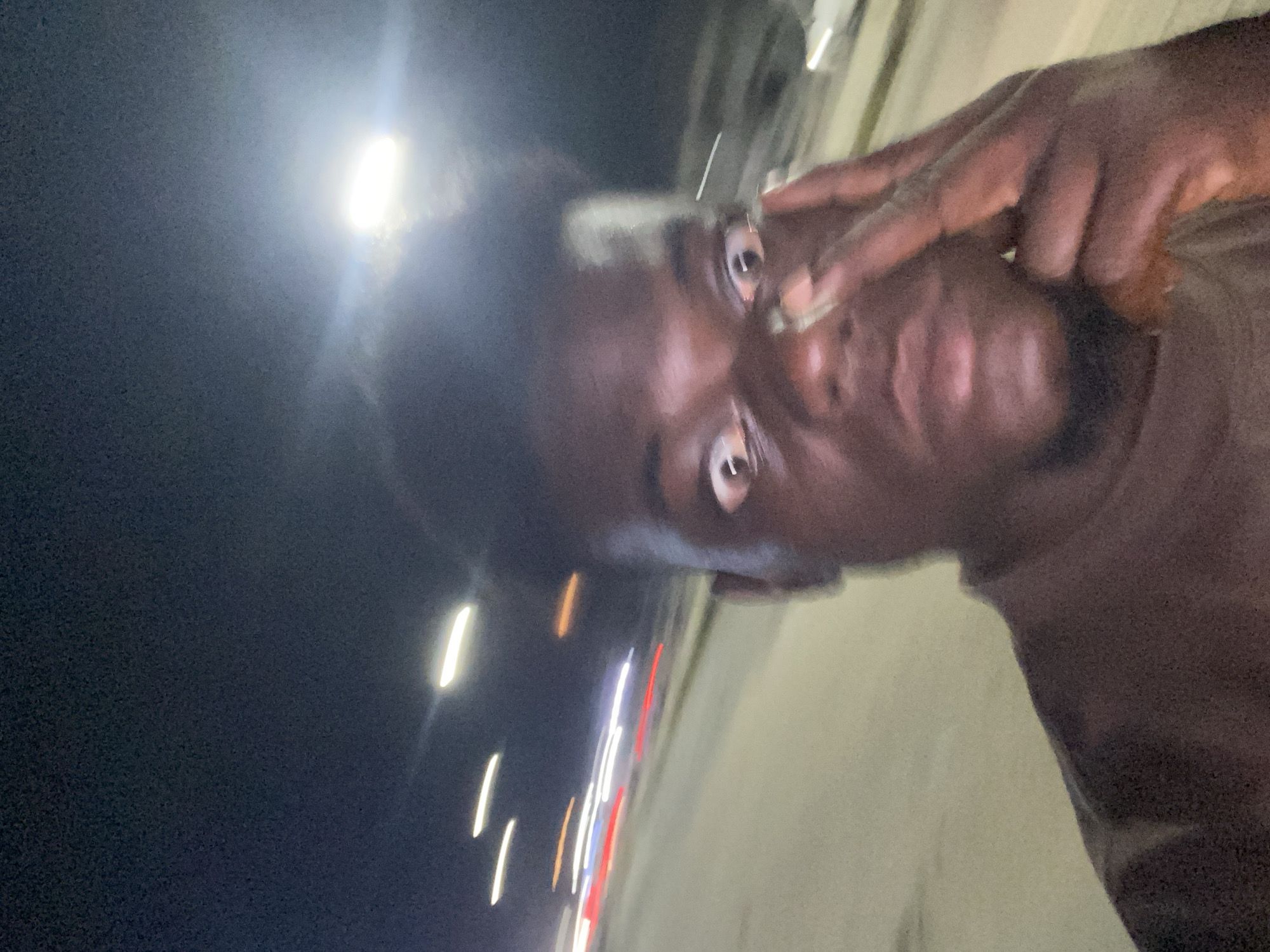 A close-up, low-angle photograph shows a dark-skinned person with short hair, holding two fingers up to their right eye, presumably creating a "V" or peace sign gesture. The individual's face is partially illuminated, revealing distinct facial features like their eyes, nose, and mouth. The background suggests a nighttime or low-light environment, with blurred streaks of light indicative of moving traffic. The scene appears to be in an urban environment. The city and state location are unknown.