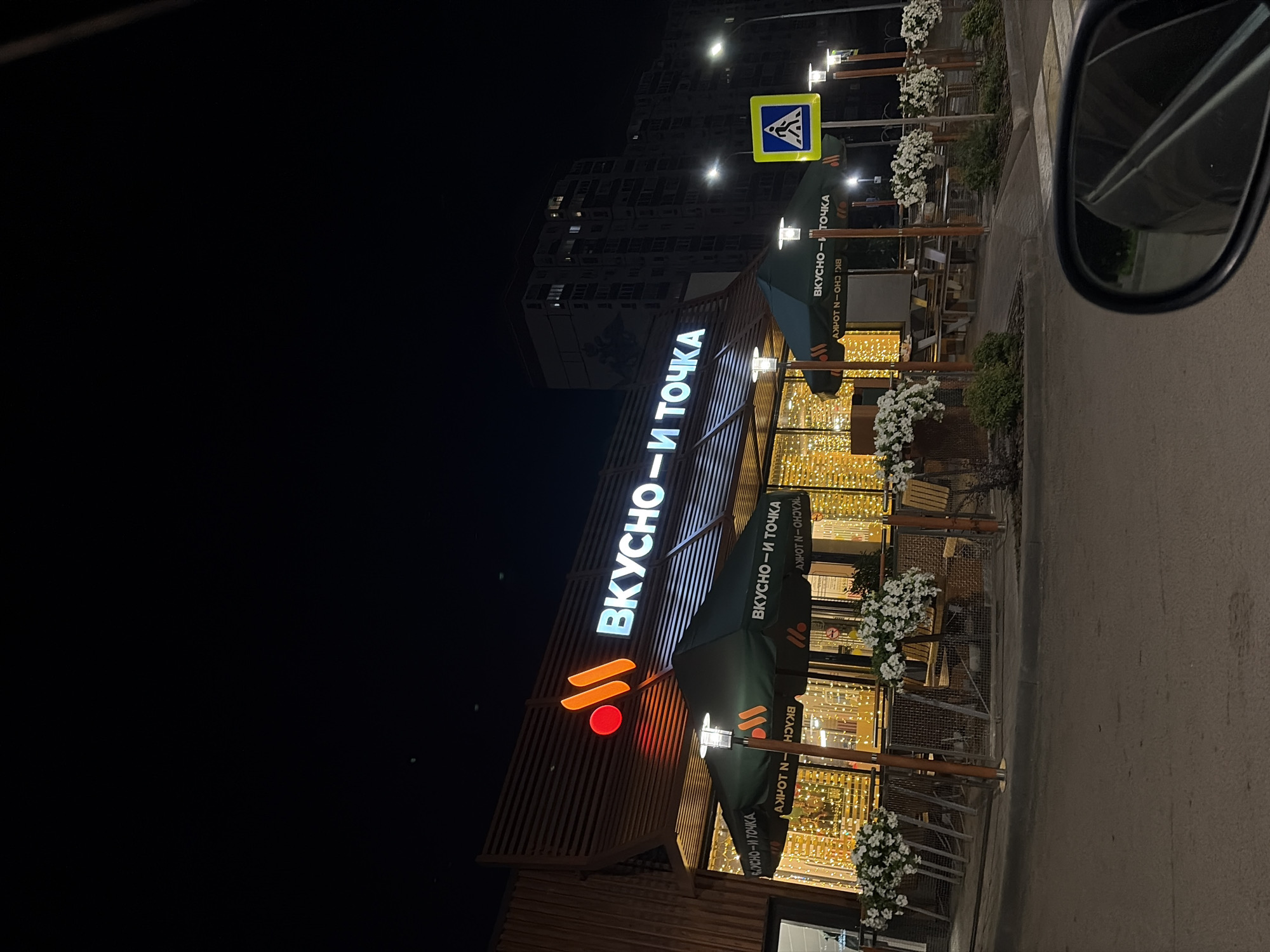 This image shows the exterior of a fast-food restaurant at night in Ufa, Russia. The restaurant has a modern design with wooden slats on the facade and illuminated signage that reads "ВКУСНО-И ТОЧКА" (Tasty-and-Point). The lights inside the restaurant are warm and inviting, and there are outdoor seating areas with umbrellas and planters filled with white flowers. A pedestrian crossing sign is visible on the left side of the image. The overall scene suggests a busy urban environment, with the restaurant being a popular spot for late-night dining. The image is taken from a car, as indicated by the side mirror in the foreground.