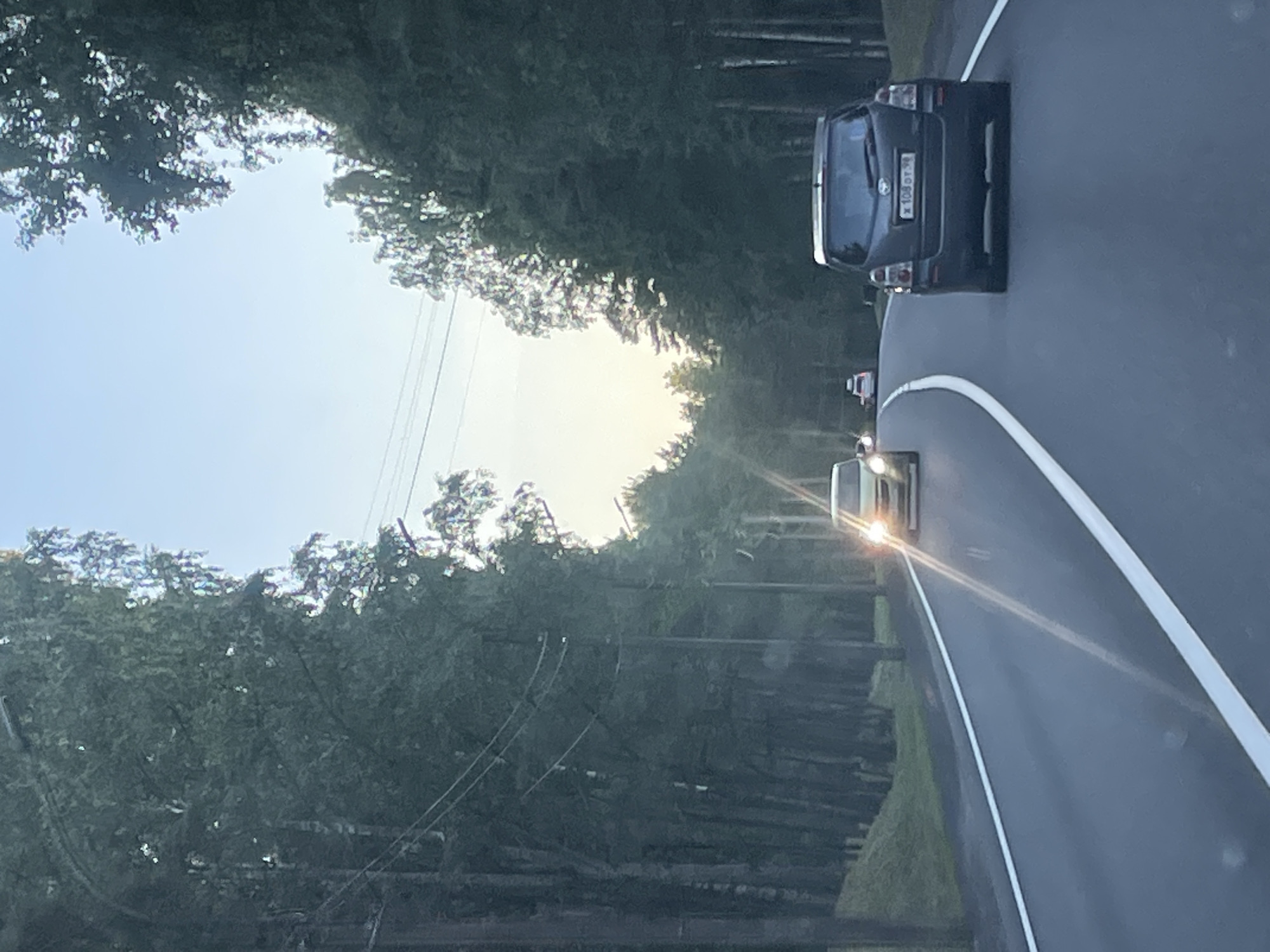 A rear view of a dark-colored SUV is captured on a paved road in Serovo, Russia. The SUV has a license plate that reads "x 108 от 98". The road is bordered by a white line on the right. On the left, a line of tall trees forms a dense forest. The sky is clear and light blue with some scattered clouds. There is another car in the distance, approaching from the opposite direction, with its headlights on. The overall lighting suggests it is either early morning or late afternoon.