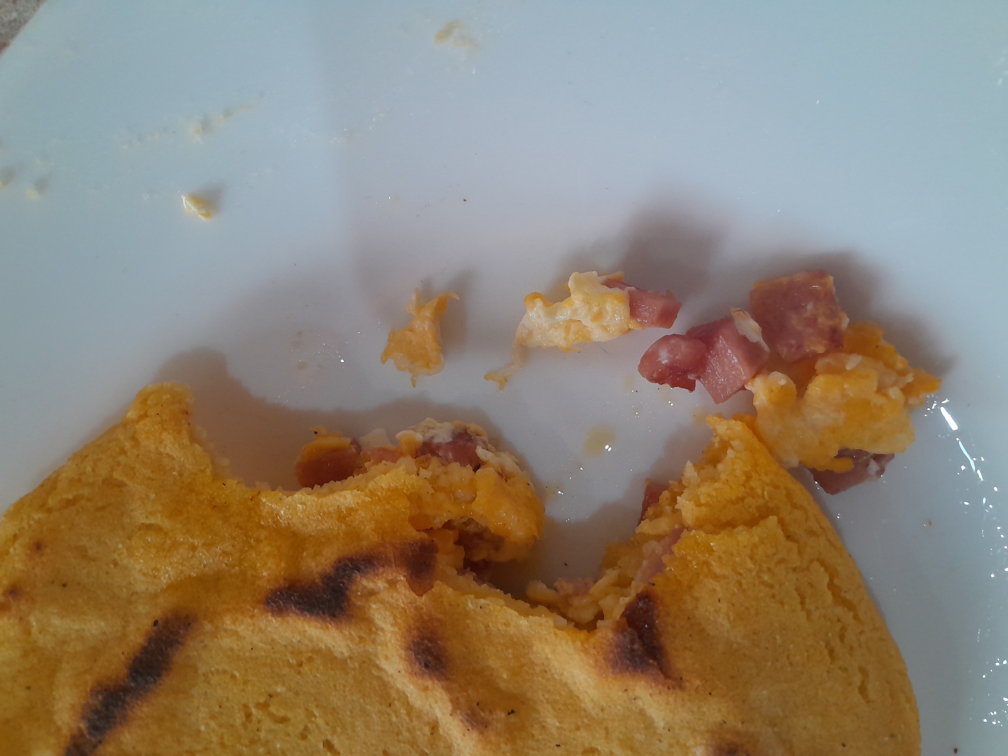 A close-up shot captures a piece of cooked corn masa with a bite taken out, revealing a filling of scrambled eggs and diced ham. The masa is golden yellow with some darker, charred spots, suggesting it was cooked on a griddle or open flame. The filling is a mixture of fluffy yellow scrambled eggs and small, pink cubes of ham. Some of the filling has spilled out onto the white surface it rests on, which appears to be a plate or placemat. The lighting is natural, casting soft shadows and highlighting the textures of the food. The scene is devoid of people or any specific environmental context beyond the food and its serving surface. There are no discernible elements that would indicate a specific location within Santa Ana, Venezuela.