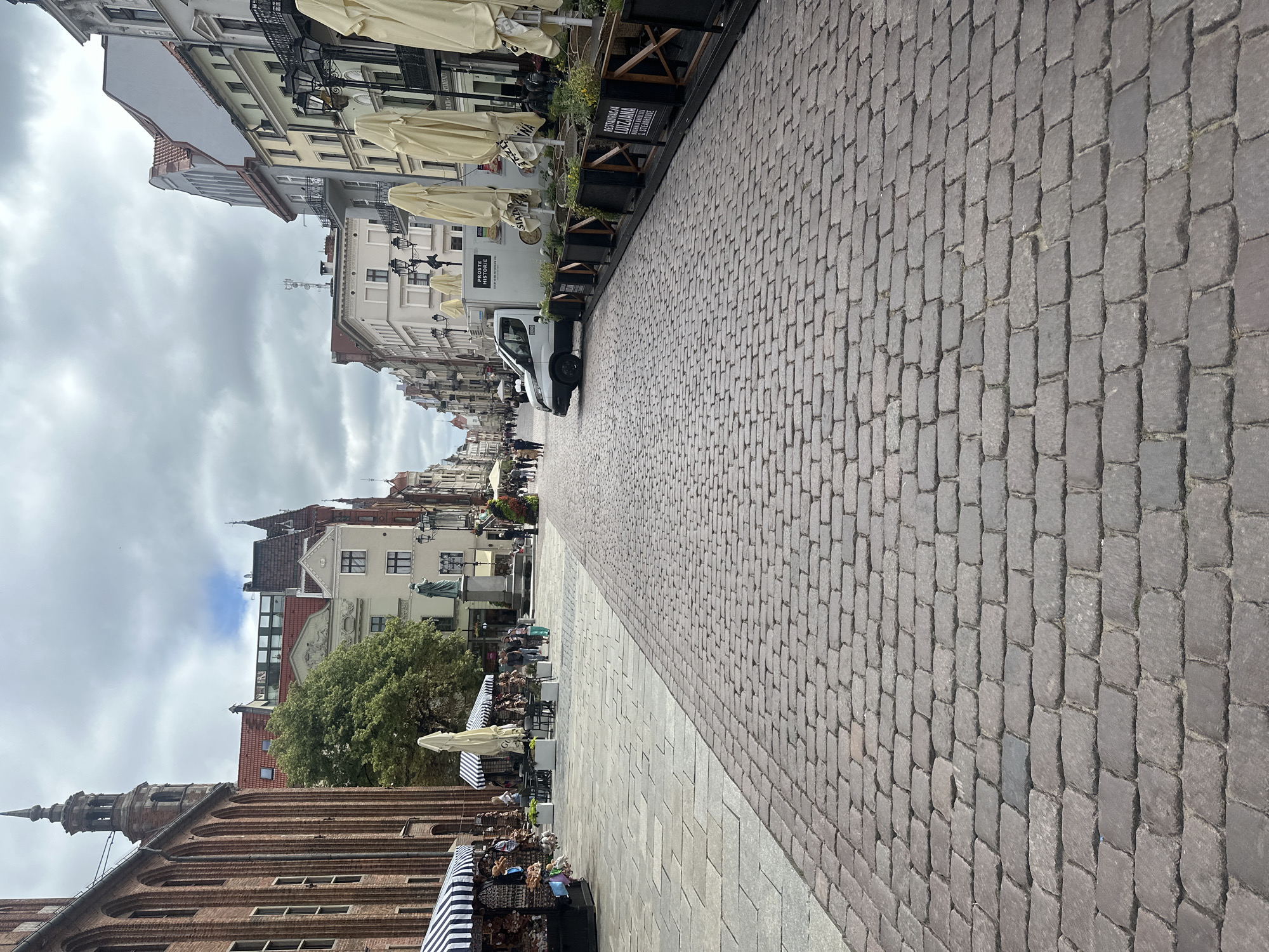 The image depicts a cobblestone street in the Old Town of Toruń, Poland, under a cloudy sky. The street is lined with historic brick and stucco buildings showcasing diverse architectural styles, including Gothic and Renaissance elements. On the left side, stalls with awnings display merchandise, and a few people are browsing. Further down the street, a statue stands in a square, surrounded by more buildings. On the right side of the street, outdoor seating areas with large umbrellas are set up in front of restaurants. A white van is parked on the street. Pedestrians are visible in the distance, walking along the street. The perspective is from ground level, looking down the street.