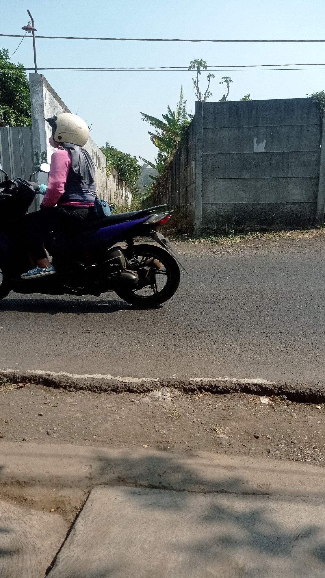 A person wearing a light-colored helmet and a pink and gray long-sleeved shirt rides a dark-colored motorcycle on a paved road. The motorcycle has a blue and black body. The person is wearing light blue shoes. A blue bag is attached to the back of the motorcycle. The road is bordered by a dirt shoulder and a concrete wall. Green foliage, including banana trees, grows near the wall. Overhead, power lines are visible against a clear blue sky. The image appears to be taken in Bedali, Indonesia, based on the context.