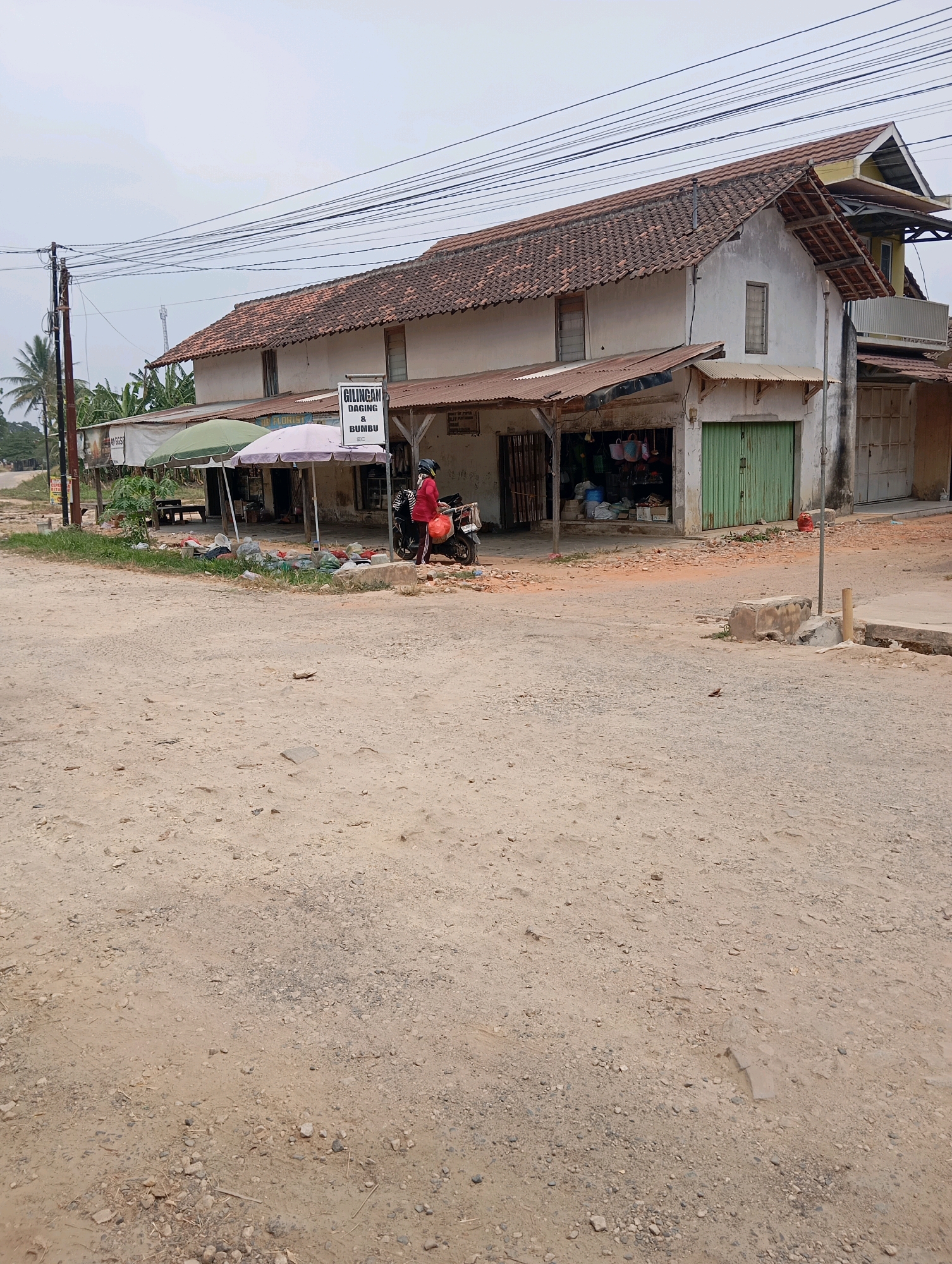 The image depicts a street scene in Batang Hari Ogan, Indonesia, featuring a roadside shop with a residential structure above. A person wearing a red shirt and a helmet is standing next to a motorcycle, holding a plastic bag. The shop is open, displaying various goods, and is covered by awnings. A sign above the shop entrance reads "GILINGAN DAGING & BUMBU". The ground is dirt and gravel, with scattered debris. Overhead power lines crisscross the sky. The overall environment appears to be rural or semi-urban.