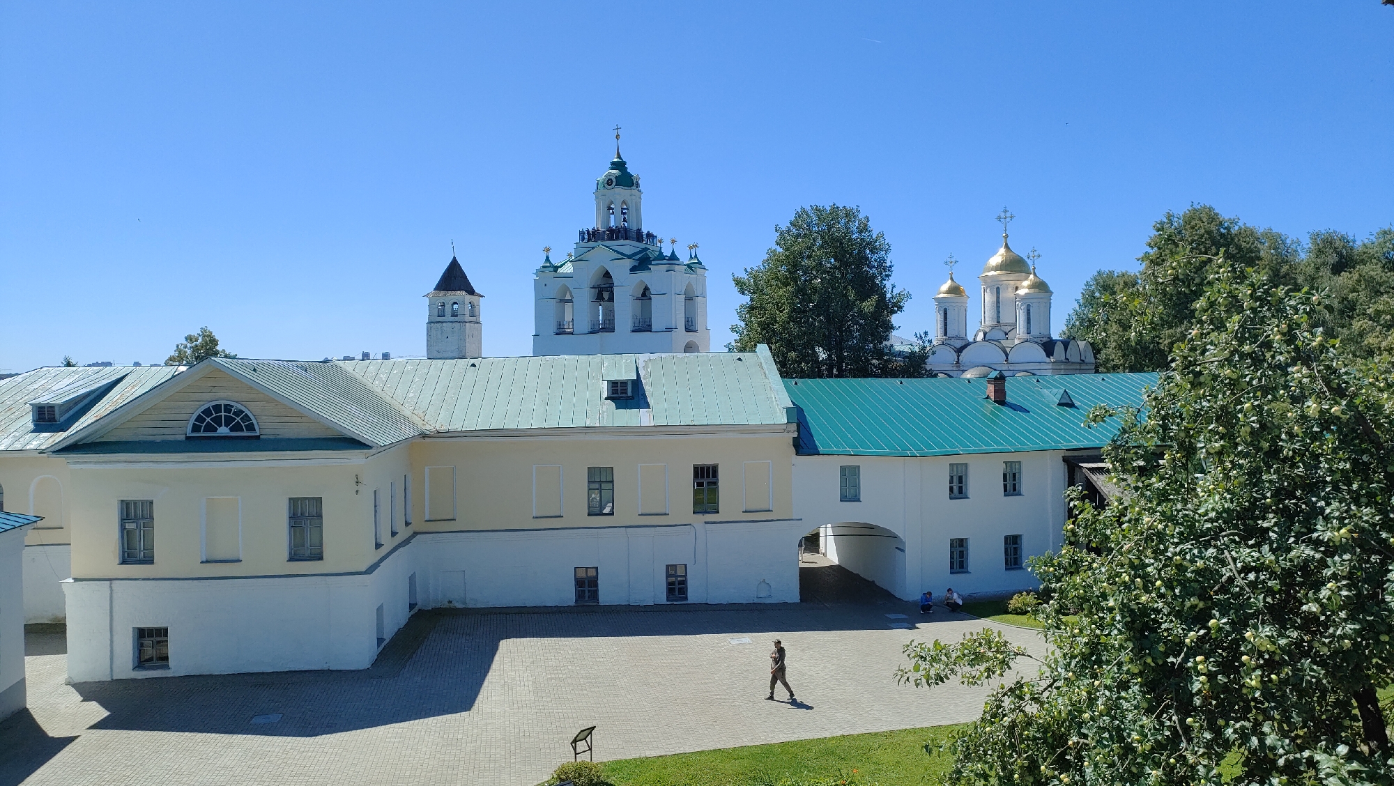 The image shows a courtyard with several buildings and religious structures under a clear blue sky. In the background, there are three prominent religious buildings. The leftmost is a white bell tower with a dark, pointed roof and arched windows. To its right is a large white bell tower with multiple levels, green accents, and numerous arched openings. The rightmost religious structure features three golden domes on white buildings, characteristic of Orthodox churches.
In the foreground, a low-rise building with a light yellow facade and a white base is visible. This building has a green metal roof with several small windows or vents. To the right of this building, another white structure with a green metal roof forms an archway, leading to another part of the complex. A person is walking across the paved courtyard in front of these buildings. The courtyard is paved with light-colored bricks. To the far right of the image, a lush green tree with visible green fruits hangs over the courtyard, partially obscuring the view of the buildings.
This scene depicts the Spaso-Preobrazhensky Monastery in Yaroslavl, Russia. The monastery is an architectural ensemble dating back to the 12th century, located on the confluence of the Volga and Kotorosl rivers. The white building with the yellow facade appears to be part of the monastic cells or administrative buildings, while the religious structures in the background are likely the Cathedral of the Transfiguration of the Savior and the bell tower. The overall atmosphere is one of a sunny day in a historic religious complex.