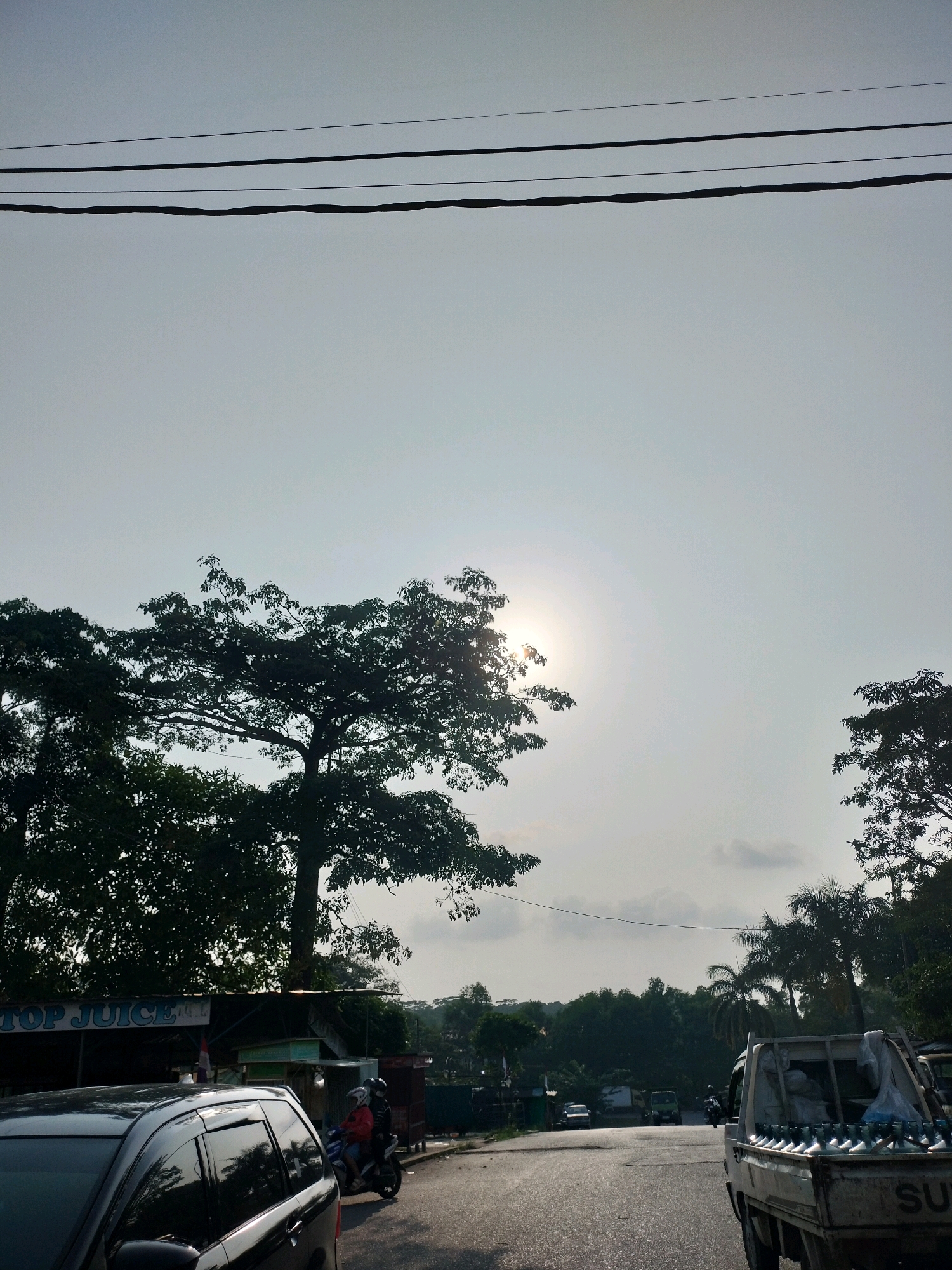 The image captures a street scene in Batam, Indonesia, likely during the late afternoon or early morning given the low angle of the sun, which is visible through the hazy sky and silhouetted by trees. The foreground is dominated by the side of a dark-colored car, its windows reflecting the surrounding environment. To the right, a white pickup truck is partially visible, carrying rows of large plastic water bottles in its bed. Further down the road, a motorcycle with two riders, one wearing a red jacket and the other a blue one, is also in motion.
The road itself appears to be asphalt and slopes slightly upwards. On the left side of the road, there is a building with a sign that reads "TOP JUICE." Several other structures and trees line the street, forming a backdrop of vegetation and buildings. Overhead, power lines stretch across the sky. The overall atmosphere is one of a typical day in a semi-urban or suburban area of Batam.