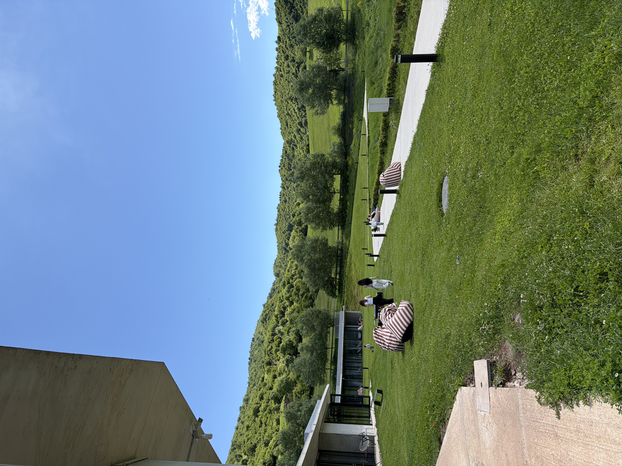 The image depicts an outdoor scene likely in Chkalov, Armenia. A grassy lawn extends from the lower right towards the center, dotted with striped beanbag chairs and some people who appear to be reclining. A white walkway runs along the right side, edged with what looks like a manicured hedge. Beyond the lawn, a hillside covered in trees and greenery rises towards the horizon. The sky is clear and blue, with a few wispy clouds near the top edge. A building is partially visible on the left side of the image, with architectural details indicating a modern or contemporary design.
