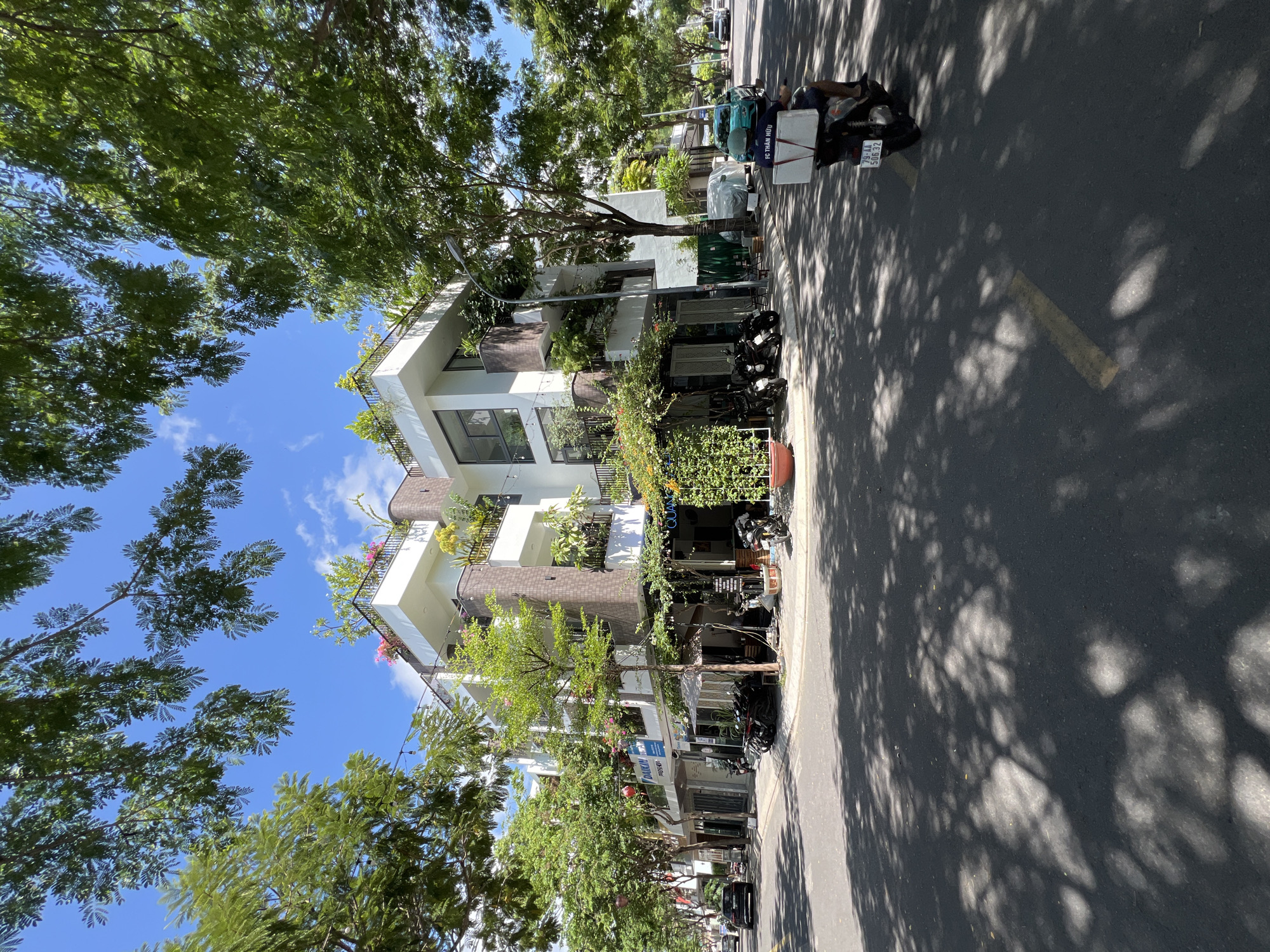 This image depicts a street scene in Nha Trang, Vietnam, on a sunny day with dappled sunlight filtering through tree canopies. In the foreground, a paved road is visible with yellow lane markings. A person wearing a helmet and a dark shirt with white lettering rides a scooter with a large white box on the back. The scooter has a license plate displaying "79-AA 50632".
Dominating the mid-ground is a multi-story modern building with white facades, balconies adorned with plants, and large windows. The building features a green living wall on one side and plants on its upper levels and rooftop. Several parked motorcycles are visible in front of the building.
Along the street, businesses with awnings are present, and trees line the sidewalk. A black car is parked on the left side of the street. The background shows more trees and a clear blue sky with scattered white clouds, indicating a pleasant weather day in the city.