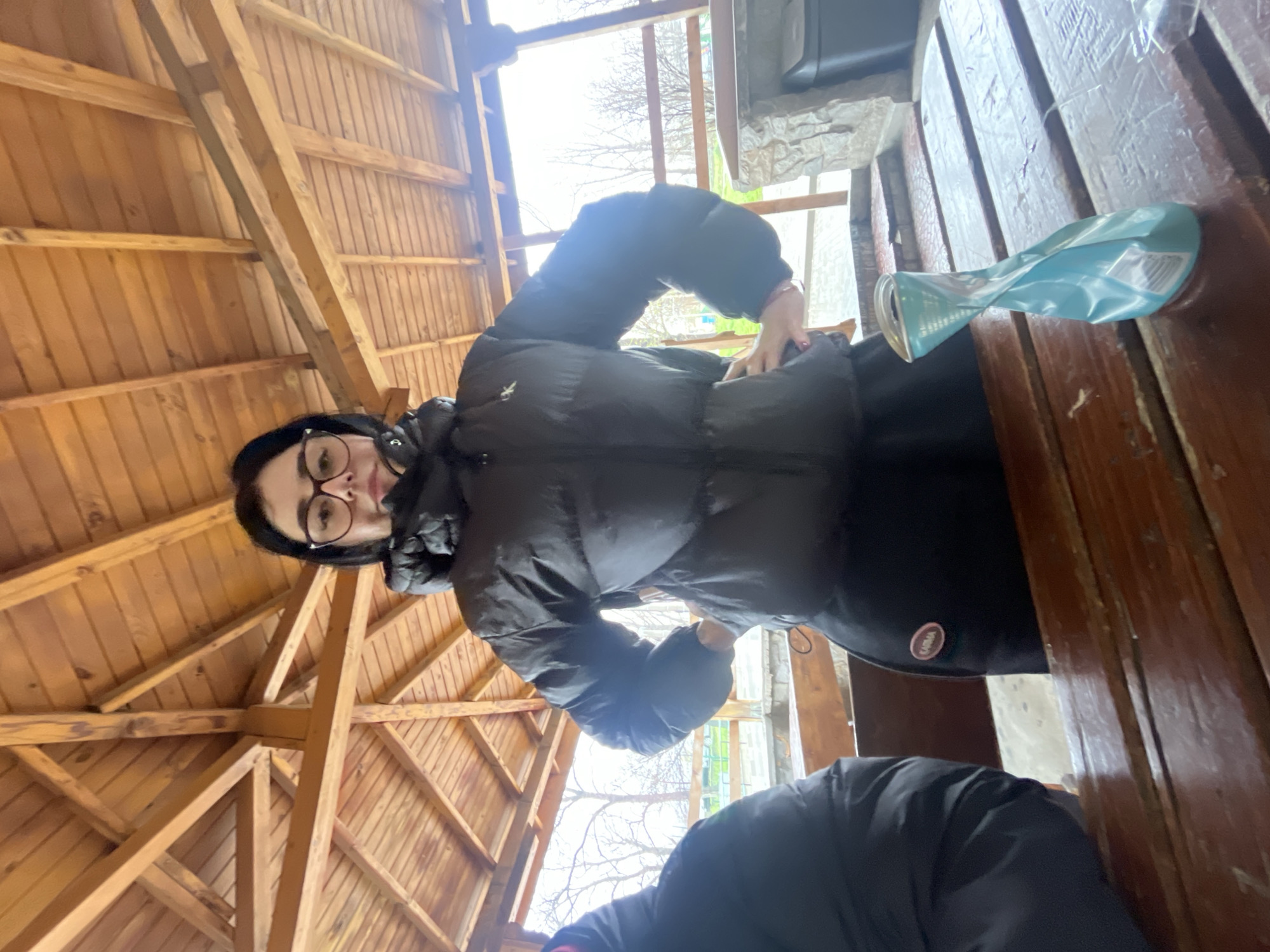 The image shows a woman with glasses wearing a black puffer jacket, sitting at a wooden table in an open-air structure with a wooden roof. A crumpled blue package sits on the table next to her. Another person wearing a black jacket is partially visible in the lower part of the image. The structure appears to be located outdoors, possibly in a wooded area. While Asenovgrad, Bulgaria is mentioned, there is no evidence or indication of this location in the image.