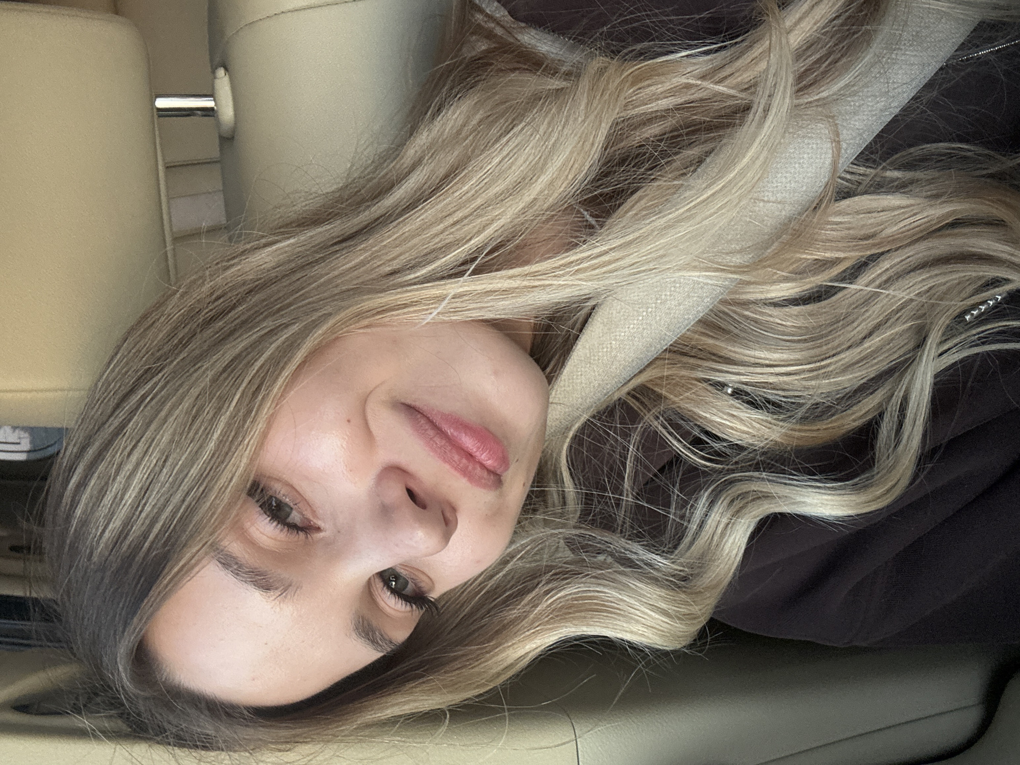 A young woman with blonde hair is in the passenger seat of a car. She is looking at the camera and smiling. The car's interior is visible, with a tan leather seat and headrest. The woman is wearing a dark brown hoodie with a light tan seatbelt across her chest. Her hair is long and wavy, with blonde highlights. The background is out of focus, but it appears to be a city street. The image is a selfie taken by the woman. There is no indication of the city of Chaikovsky, Russia, in the image.