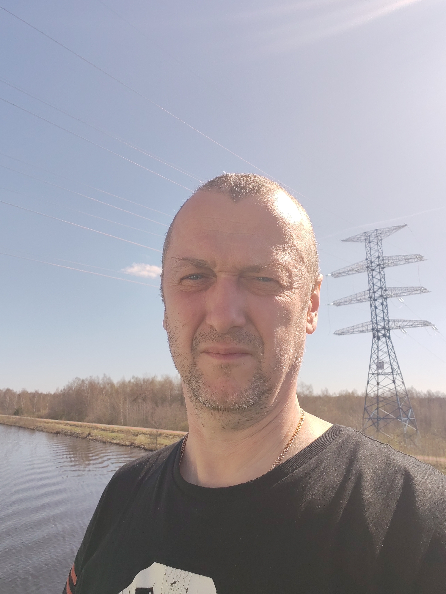 A selfie of a middle-aged man is taken outdoors on a sunny day. The man is positioned in the foreground, wearing a black t-shirt and a gold chain around his neck. He has short, light-colored hair, and stubble on his face.
Behind him, a body of water stretches into the distance, with a grassy bank and bare trees lining the opposite shore. An electrical transmission tower stands prominently in the background to the right of the man, with power lines stretching across the sky. The sky is clear and blue. The location is Unknown, Unknown.