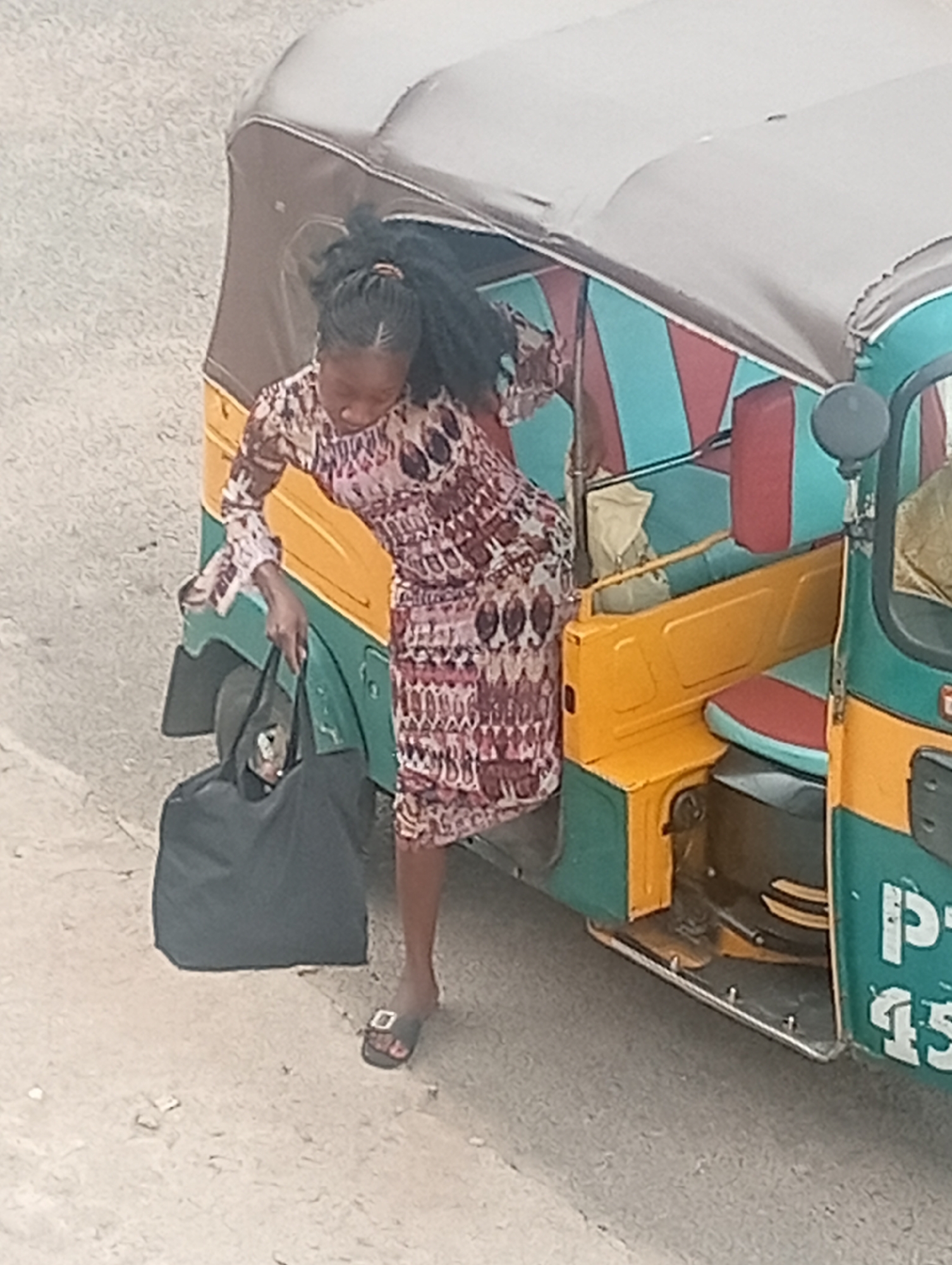 A woman is exiting a vehicle in what is likely Barakin Akawo, Nigeria. The vehicle is a three-wheeled passenger vehicle, commonly known as a "keke" or "tricycle". It is painted in a combination of colors including green, yellow, and teal with red accents on the seating area. The woman is wearing a patterned dress, slides and has a large black bag. She is in the process of stepping out of the keke. The background shows a concrete or paved surface. The image quality is grainy, with a slight blur.