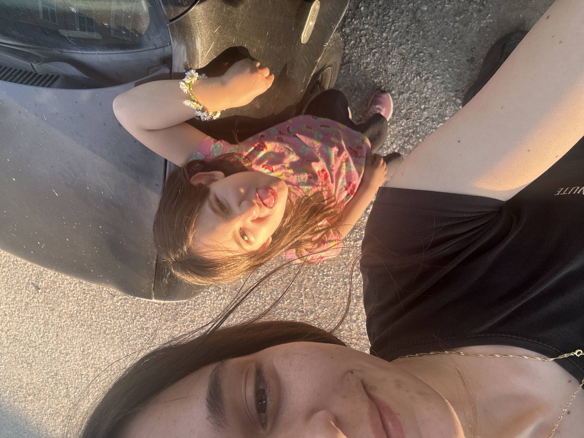 The image appears to be a selfie taken by an adult with a child. The adult's face is visible in the lower portion of the frame, showing dark hair and a gold necklace. The child is positioned above, lying sideways, with a pink shirt and a daisy bracelet on their wrist. The child is sticking out their tongue, making a playful expression. A portion of a black car is visible next to the child. The background appears to be a paved surface. Given the lighting conditions, it seems to be taken outdoors during daylight. There is no specific evidence in the image to confirm the location is Resen, Bulgaria.