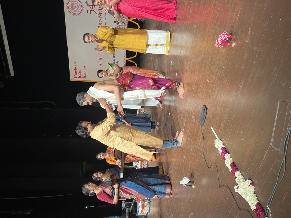 The image shows a stage in the Pracheen Kala Kendra, located in Chandigarh, India (latitude: 30.733, longitude: 76.79). There are six people on stage. A man in a gold shirt is draping a white shawl over the shoulders of another man wearing a pink and gold outfit. They are standing in the center of the stage. To the left, there are three women standing, one of them holding a certificate. Another man, wearing a yellow kurta, is standing to the right clapping his hands. A woman in a red saree is standing on the far right. On the stage, there is a floral garland and two red microphone stands. The backdrop behind the people reads “Pracheen Kala Kendra, All India 54th Rao Nritya. The event appears to be an award ceremony.