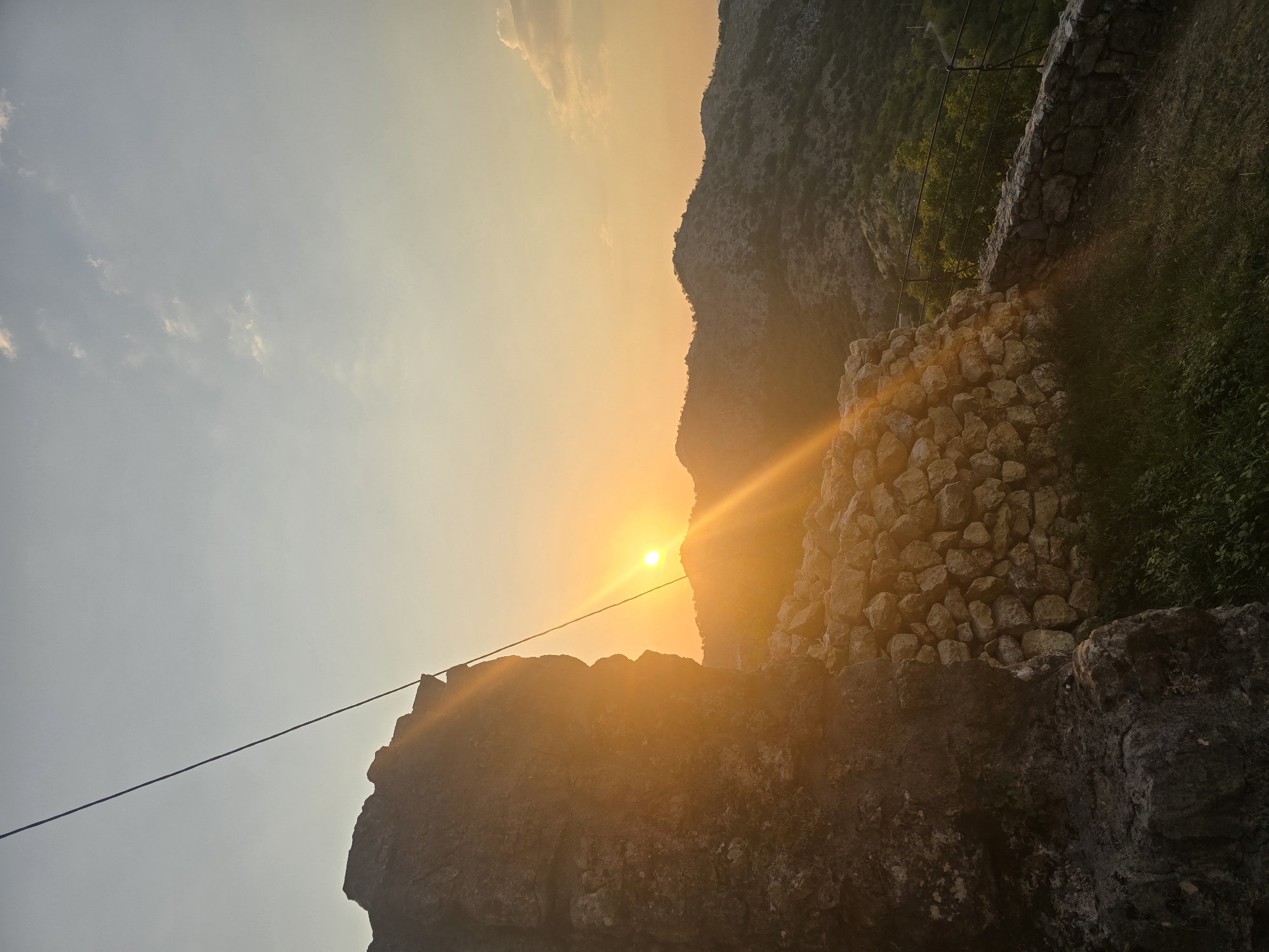 The image depicts a sunset scene from a hilltop overlooking a mountainous landscape. In the foreground, a large, weathered rock formation stands on the left, partially obscuring a stacked stone wall. A thin wire or cable stretches diagonally across the upper left portion of the frame. The setting sun, a bright orange orb, is positioned just above the distant mountain ridge, casting long rays of golden light across the scene and illuminating the clouds in the sky. The mountains in the background are covered with sparse vegetation. The overall atmosphere is serene and natural, with no people or animals visible. The location is identified as Gornja Vrbica, Montenegro.