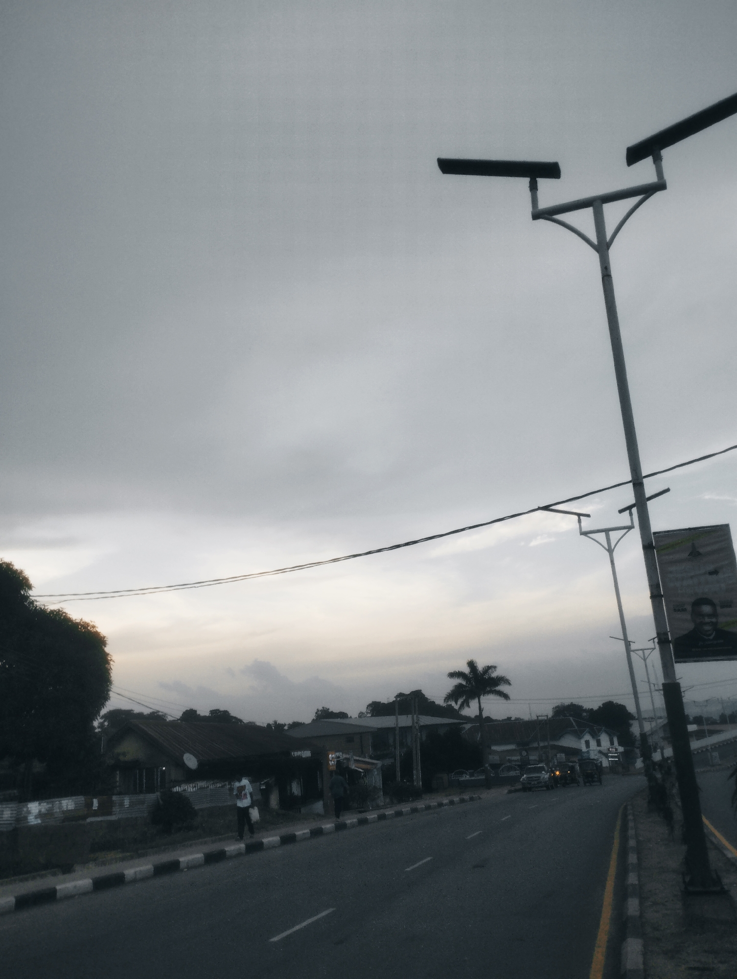 The image depicts a street scene in Jos, Nigeria, under an overcast sky, possibly during dawn or dusk. A paved road with visible white lines runs through the center of the frame, bordered by a sidewalk with black and white markings. To the left of the road, there are buildings with dark roofs and some trees, including a prominent palm tree in the distance. Streetlights with modern designs are visible, positioned along the road. A billboard featuring a portrait of a man is also present on the right side of the road. Several vehicles, including cars and what appears to be a tricycle, are on the road. Two individuals are visible walking on the sidewalk. The overall atmosphere is subdued, characterized by the gray sky and the muted tones of the urban environment.