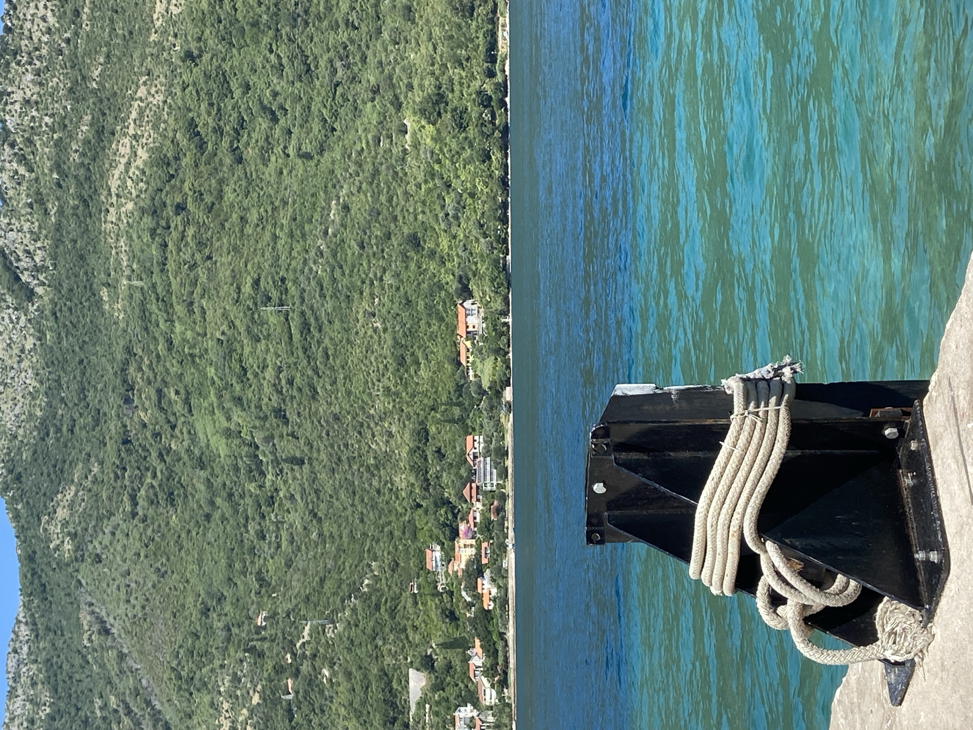 The image depicts a scene in Lepetane, Montenegro, on a sunny day. In the foreground, a thick, beige rope is coiled and secured to a black metal bollard on a concrete dock. The bollard is bolted to a concrete base. Behind the bollard, the turquoise water of the bay is calm, with slight ripples. In the background, a shoreline is lined with trees and several houses with red-tiled roofs. Beyond the shoreline, a steep, densely vegetated green mountain rises sharply. The sky above is clear and blue.
