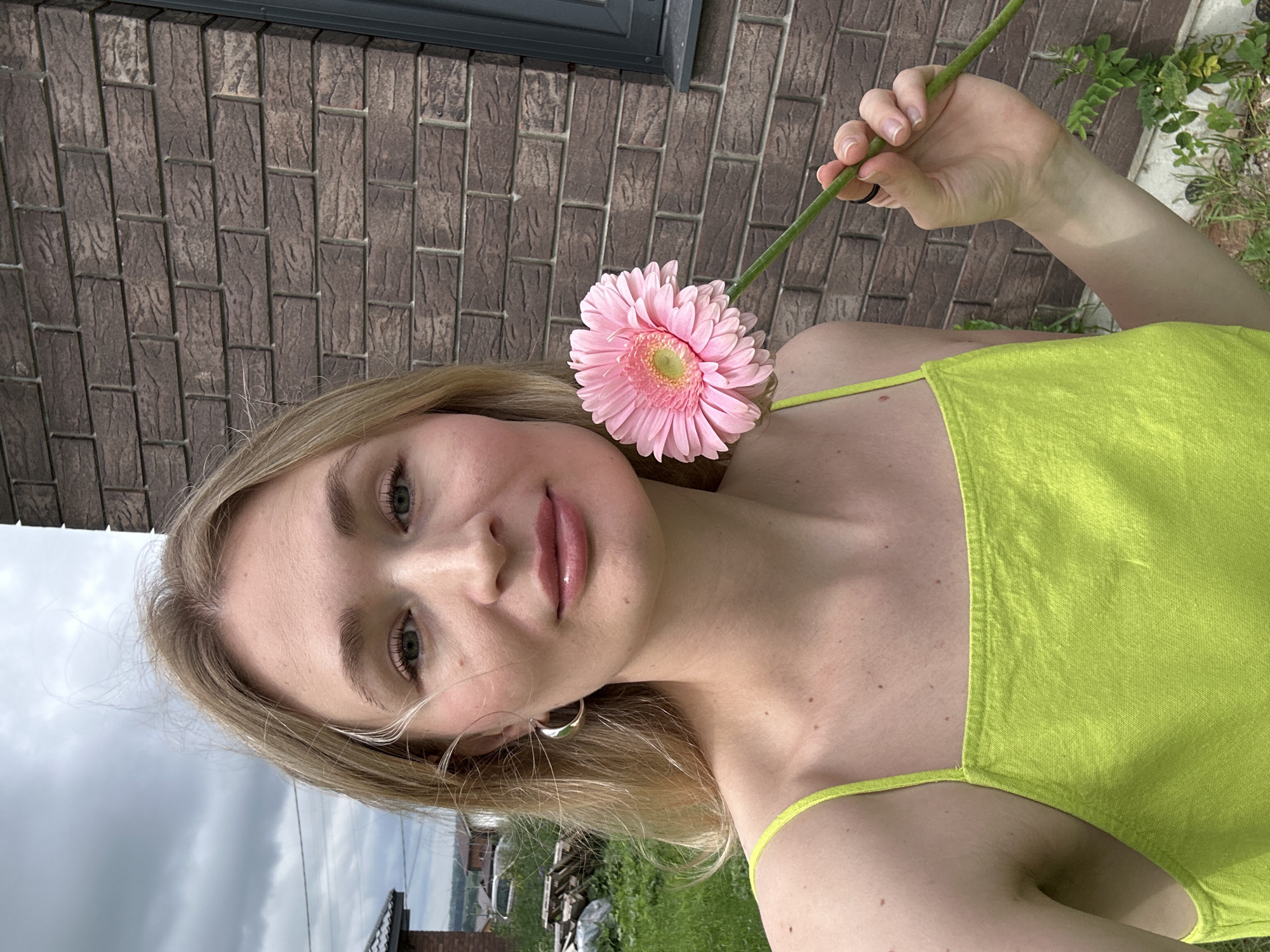 A young woman with blonde hair and green eyes, wearing a bright green tank top, holds a pink gerbera daisy in her right hand in front of a brick wall. She is outdoors, with green grass and trees in the background. The sky is overcast. The woman is looking towards the camera. The location is in the city of Mokino, Russia.