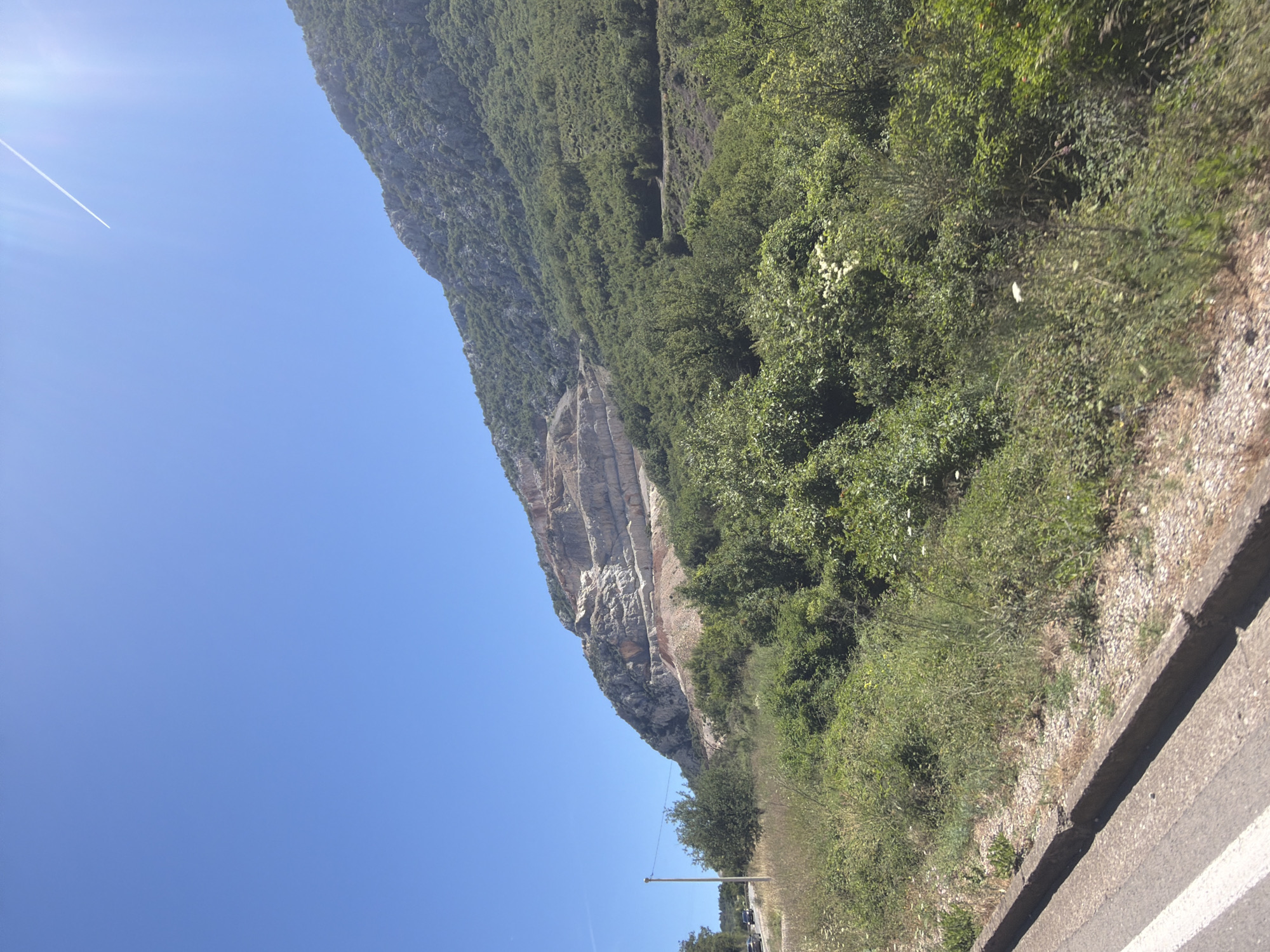The image depicts a sunny day in Mišići, Montenegro, with a clear blue sky and a bright sun. The foreground shows a road with a white line on the left side, bordered by a concrete curb and gravel. Beyond the road, dense green vegetation, including trees and bushes, covers the lower slopes of a mountain. The mountain itself features a large, excavated quarry with distinct horizontal layers, revealing exposed rock faces. A single utility pole stands to the left of the road. In the distance, a thin white contrail stretches across the sky, indicating the presence of an airplane. The overall scene is that of a natural landscape impacted by industrial quarrying, under a bright, clear sky.