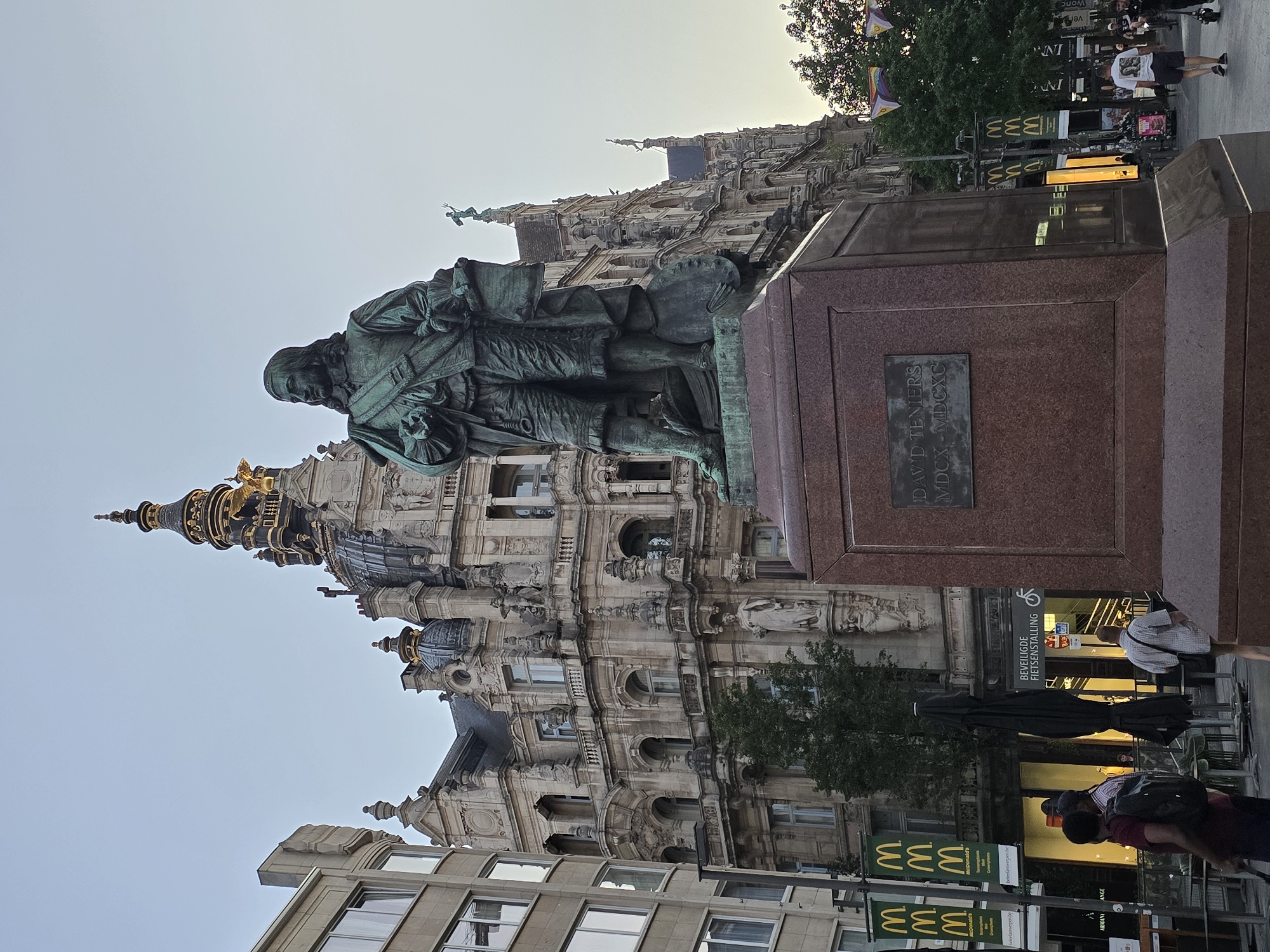 The image shows a bronze statue of a man in historical dress, standing on a large, reddish-brown stone pedestal. The statue depicts a man with long hair and a serious expression, holding a scroll in his left hand and a palette in his right. The pedestal has a plaque inscribed with "DAVID TENIERS MDCX - MD M MGCXC". Behind the statue is an ornate, multi-story building with classical architecture, featuring intricate stonework, arched windows, and decorative domes. The building is a prominent landmark. In the foreground and background, there are elements of a city street scene. McDonald's banners are visible, along with people walking and standing. Some trees are present, and there is a streetlight. The sky is a pale blue, indicating either early morning or late evening. This scene is located in Antwerp, Belgium, specifically at the Meir square, home to the statue of the famous Flemish Baroque painter David Teniers.
