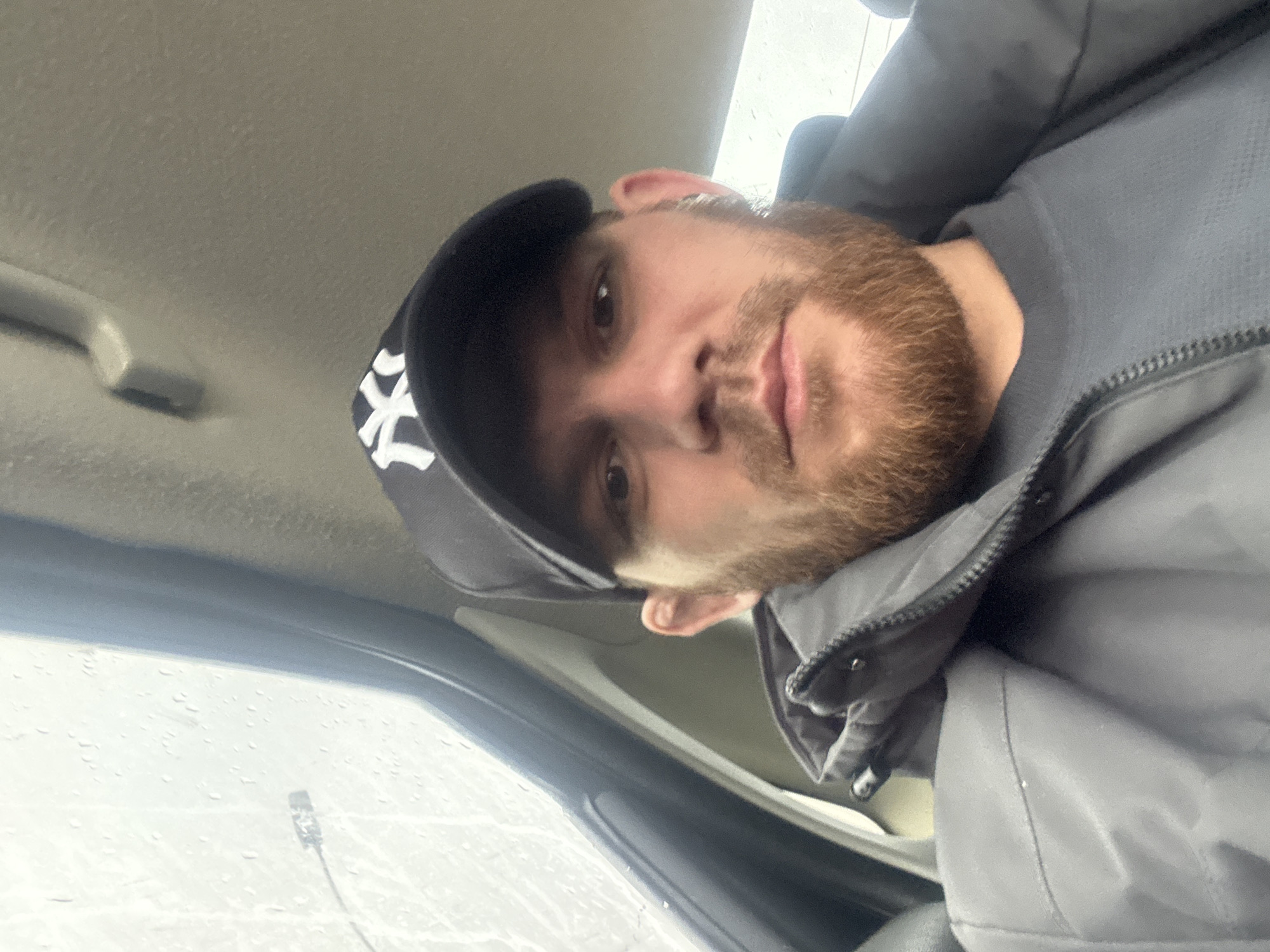 A man with a reddish-brown beard and dark hair is depicted from the chest up, positioned inside a vehicle. He is wearing a dark grey or black baseball cap with a prominent white "NY" logo on the front. Over a darker inner layer, he wears a heavy, dark grey zippered jacket with a high collar. His head is tilted, and he looks directly at the camera with a neutral to slightly thoughtful expression.
The setting appears to be the interior of a car or similar vehicle, with a light-colored headliner visible above him and parts of the car's interior, including a grab handle on the left and window frame on the right. Through the window, raindrops are clearly visible, indicating rainy weather. The view outside is bright but diffused, suggesting an overcast day. A faint outline of a distant structure or pole can be seen through the wet window. The overall lighting is soft and natural, consistent with daytime.
The activity appears to be a selfie or a close-up portrait taken within the vehicle, possibly while parked or waiting. The visible text is the "NY" logo on his cap. The location context provided is Perm, Russia.