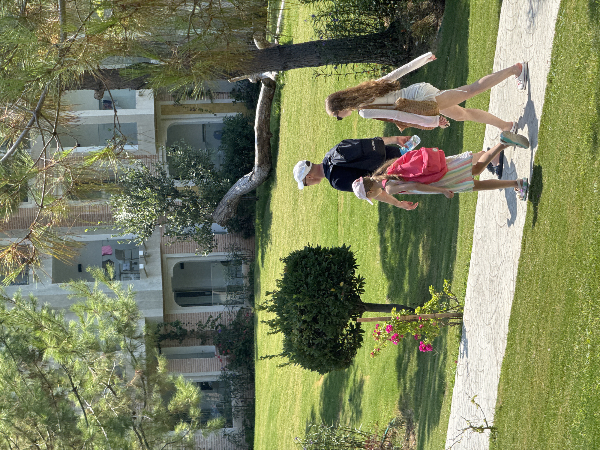 A family of three, including two young girls and one adult male, walks along a paved path in a grassy, treed area in front of a multi-story building. The man, wearing a black t-shirt, white cap, and backpack, walks slightly behind the two girls. One girl, in a striped dress and pink backpack, walks ahead of him. The other girl, with long blonde curly hair, wears a white dress and carries a woven bag over her shoulder. They appear to be on vacation in Fethiye, Turkey. The building has balconies with railings, and some clothes are hanging on a balcony on the second floor. The ground is covered in lush green grass, and trees with pine needles are visible throughout the scene. A small, trimmed bush stands in the middle of the lawn, with a flowering pink plant next to it. A large, fallen tree trunk lies horizontally on the grass in the background.