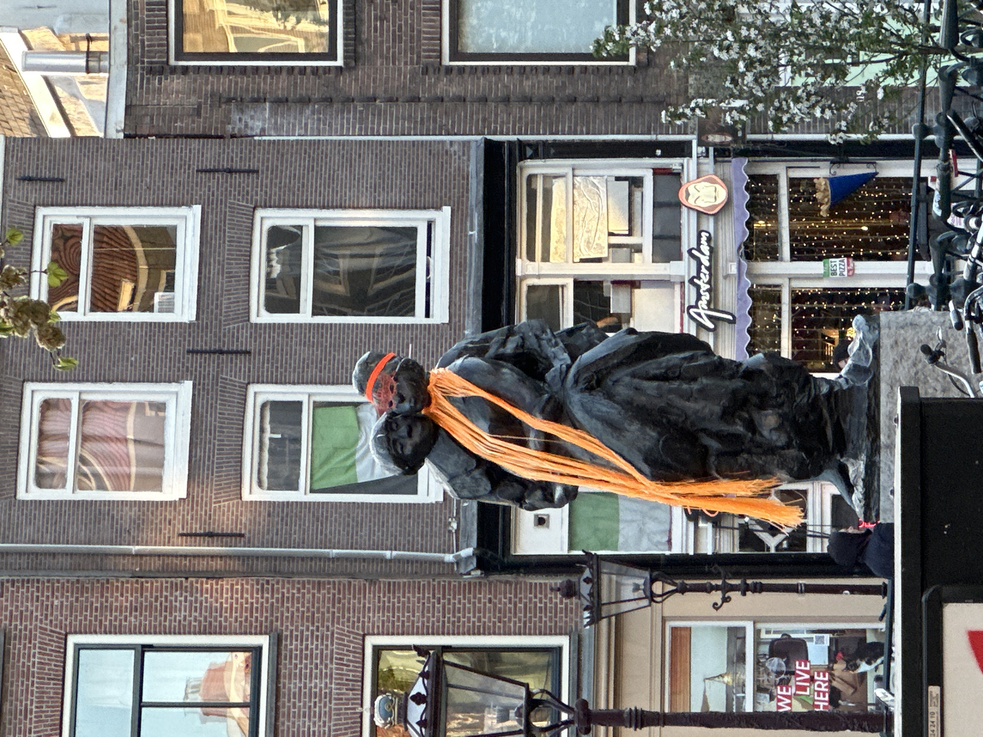 A statue of Anne Frank and her father, Otto Frank, with orange ribbons, stands along a canal in Amsterdam, Netherlands, in front of a building with a brick façade and windows. Several windows reflect the buildings across the canal. In the background is the Amsterdam Pizza restaurant.