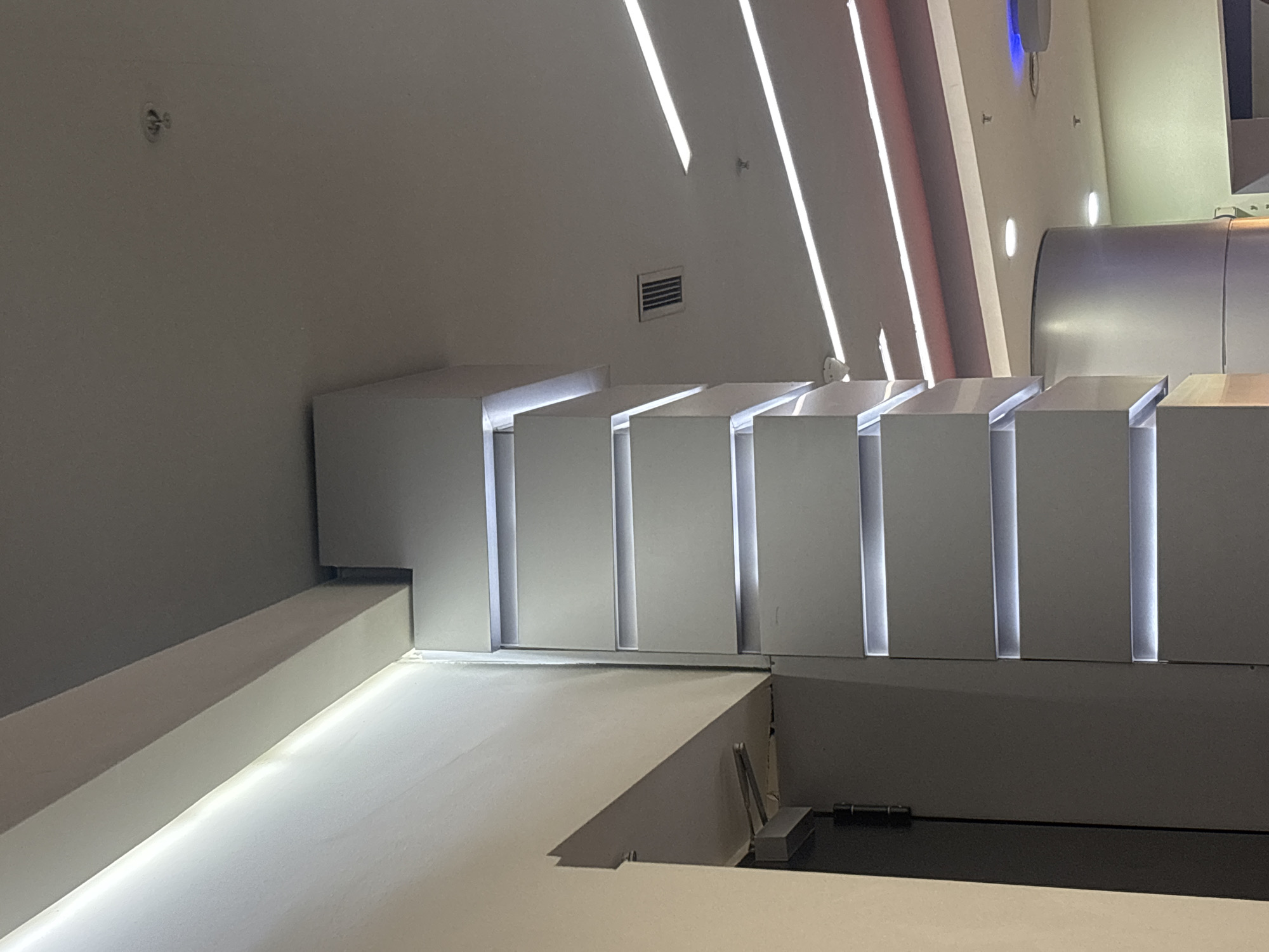 This image captures a modern, minimalist interior space characterized by sleek geometric forms and integrated lighting. The scene is devoid of people and text.
The primary subjects are architectural elements, predominantly in white and grey tones. On the left, a long, low white structure, possibly a ramp or floor section, extends into the foreground, featuring a thin strip of light along its inner edge. Above this, a white wall meets a series of tiered, rectangular structures that project horizontally from the wall. These structures are illuminated from within, creating vertical lines of soft light where their faces meet.
Further into the background and to the right, a large, curved, metallic-looking structure suggests industrial design or a futuristic installation. The walls and ceiling are also white or light grey, with several linear strip lights mounted diagonally, casting bright, narrow beams across the surfaces. A rectangular ventilation grate is visible on a central wall section. Other small, circular light fixtures are dotted on the walls in various places, and a faint blue light emanates from a fixture higher up.
The overall aesthetic is contemporary and clean, emphasizing lines, light, and form. The lighting is a crucial design element, contributing to a sophisticated and possibly futuristic ambiance. The space appears to be an interior of a public building, perhaps a gallery, museum, or corporate space, designed to be visually engaging through its architecture and illumination. No specific activity is taking place; the scene is static, showcasing the design of the environment.