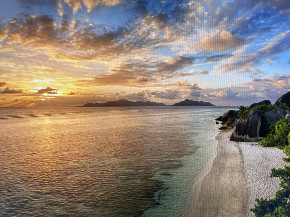 This image is a photograph of a digital display screen, presenting a natural coastal landscape under sunset or sunrise conditions. The scene is dominated by a vast expanse of calm ocean, reflecting vibrant golden and orange light, extending from the mid-ground towards a distant horizon. A low sun illuminates the right side of the frame, casting a warm glow across a sky filled with scattered clouds, ranging from intense orange and yellow near the horizon to lighter blue and white further overhead. A sandy beach occupies the foreground, curving along the lower left portion of the image, where it meets the water. A dark, rocky cliff, partially covered with green vegetation, protrudes into the scene from the bottom left. Two extremely small, indistinct, dark figures are discernible atop this rocky outcrop. The entire depiction exhibits distinct horizontal and vertical pixel grid patterns, characteristic of a digital screen capture. A faint, dark, horizontal line artifact is visible near the upper quarter of the displayed image, extending across the sky and ocean. The depicted location is a natural coastal environment, specifically Unknown, Unknown.
