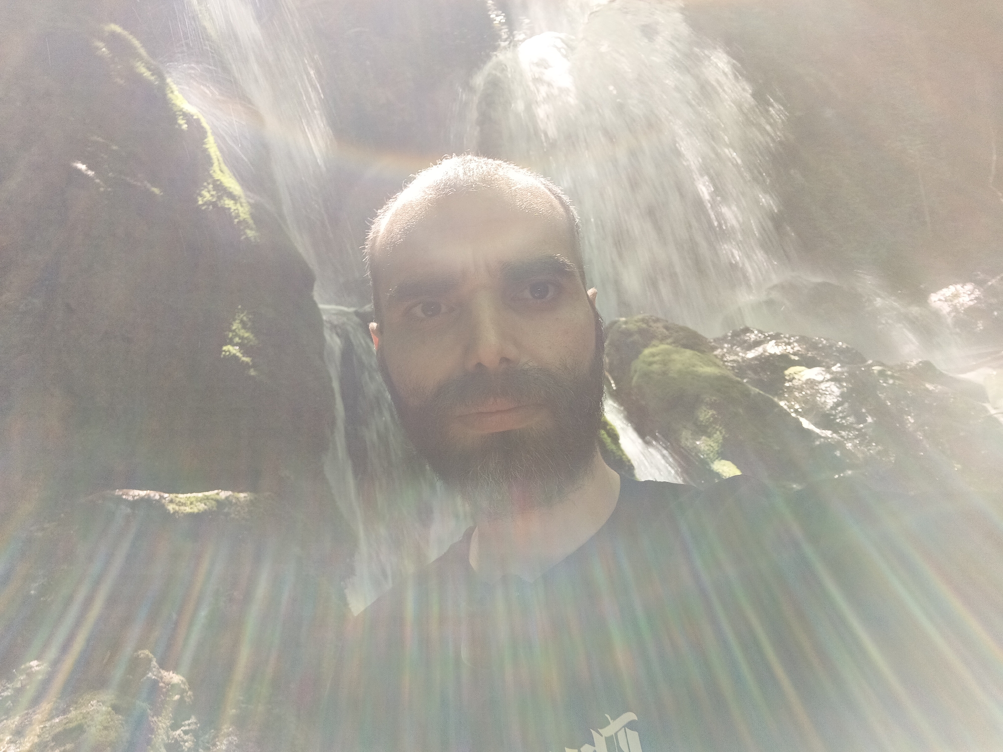 A man is taking a selfie in front of a waterfall. The waterfall is surrounded by rocks covered in moss. The man has a beard and is wearing a black shirt. The lighting is bright, creating lens flare. The waterfall is located in the city of Bachkovo, Bulgaria.