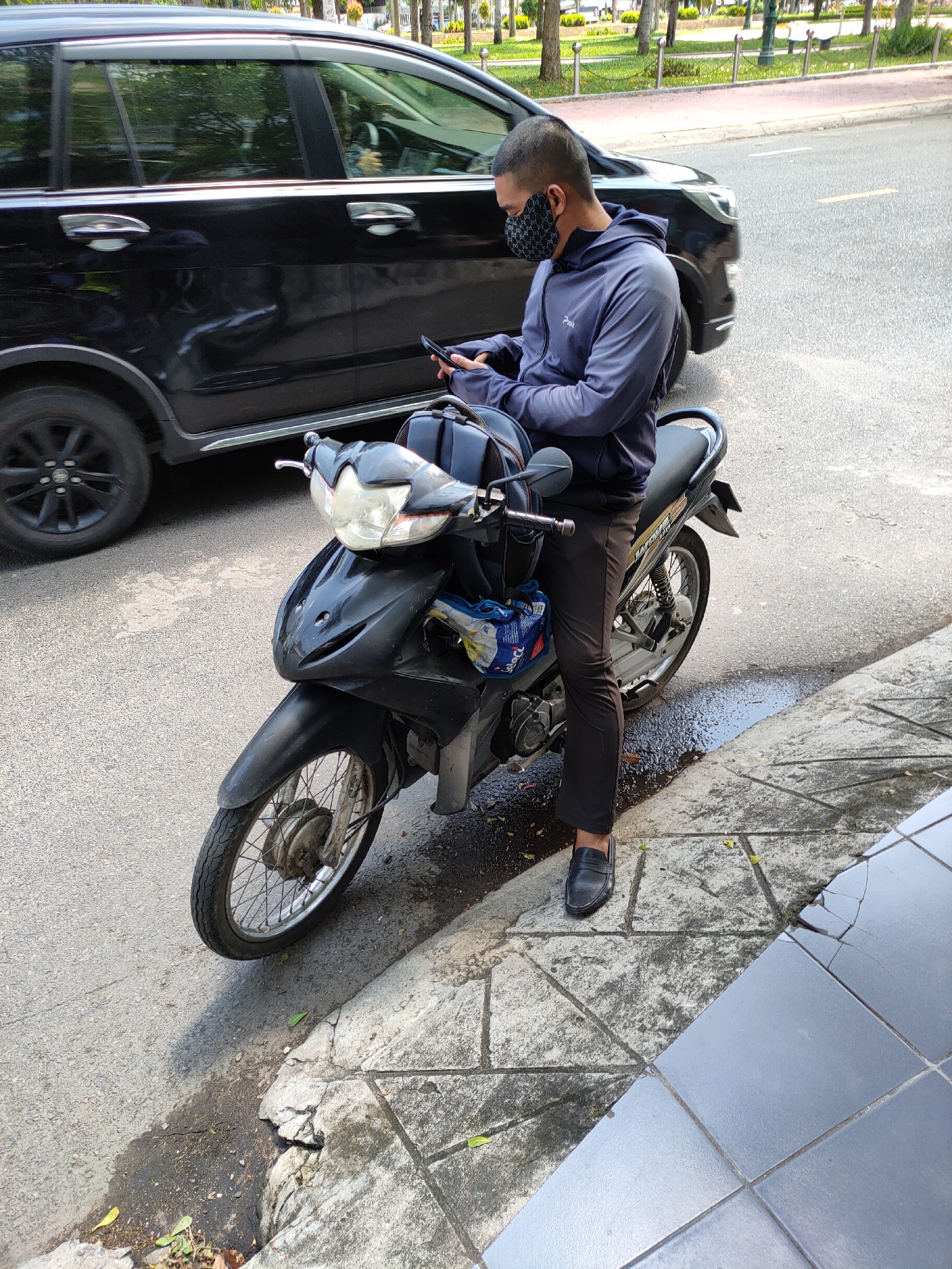 In Phan Thiết, Vietnam, a person wearing a dark face mask and a grey hooded jacket is seated on a black motorcycle. The person is looking down at a smartphone in their hands, which are resting on the handlebars. A dark-colored SUV is parked to the left of the motorcycle on a paved road. The ground next to the road is a sidewalk with cracked tiles and some greenery. The overall scene suggests a moment of pause or communication while on the go in an urban setting.