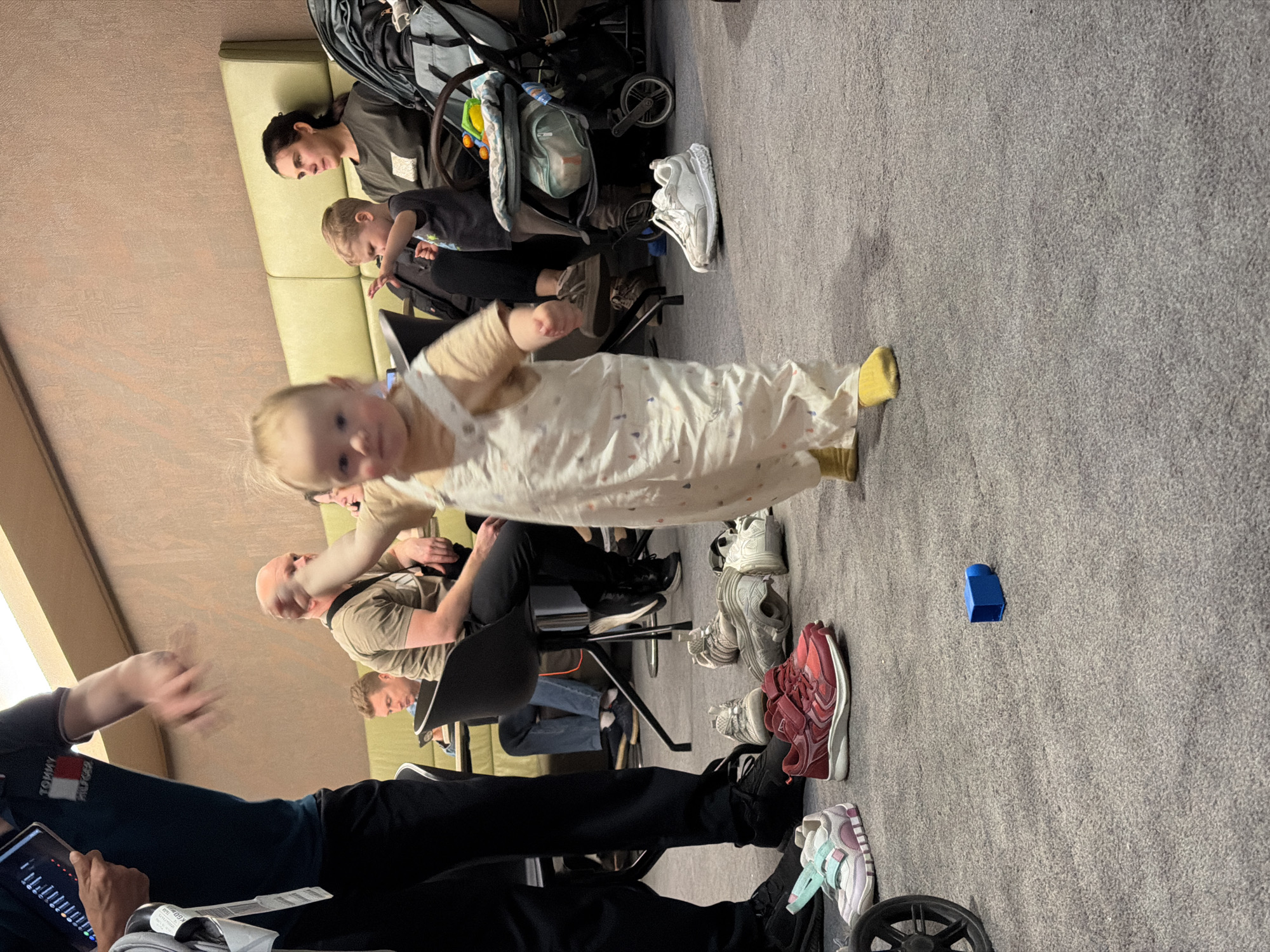 The image depicts a group of people, including adults and children, in what appears to be an indoor setting, possibly a waiting area or a community space. In the foreground, a baby wearing a light-colored outfit is in motion, with one leg extended and yellow footwear visible. The baby's face is blurred due to movement. Behind the baby, an adult male is seated, holding a tablet displaying charts and graphs. To the left of this man, another person, also seated, is visible.
Further back, a woman is sitting on a light-colored couch, looking downwards. Next to her, a young child is standing and looking in the same direction. A stroller is positioned behind them. Several pairs of shoes are scattered on the gray carpeted floor, including white, gray, and red sneakers. A small blue object is also visible on the floor. The lighting suggests it could be daytime, with natural light possibly coming from a window, though not directly visible. The scene conveys a sense of everyday life, possibly a moment of waiting or a casual gathering. There is no indication of specific weather conditions.
Visible text includes "TOMMY HILFIGER" on the t-shirt of the person holding the tablet. A label on a document near the tablet also shows some text, but it is largely unreadable. The location context provided is Khimki, Russia.