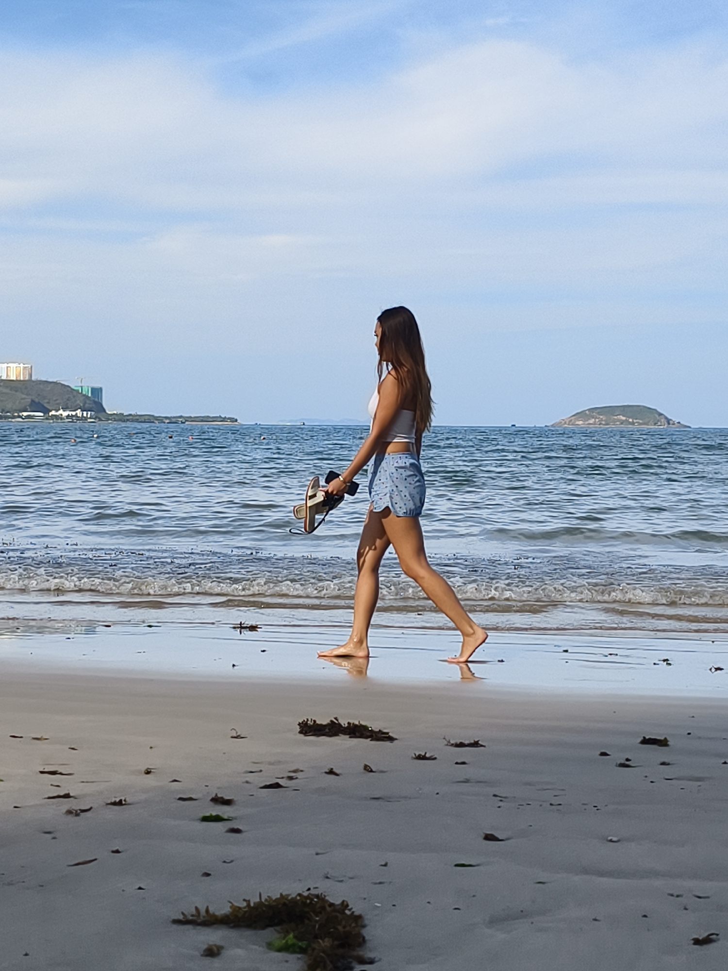 A woman walks barefoot along a sandy beach, carrying sandals in her left hand. She is wearing a white top and patterned blue shorts. The beach stretches towards the horizon, meeting the ocean. In the background, there is a city skyline and a small island offshore. Seaweed and debris dot the wet sand in the foreground. The scene is a beach setting, likely in or around the Bac Nha Trang Ward, Vietnam, based on the coastal setting and architecture.