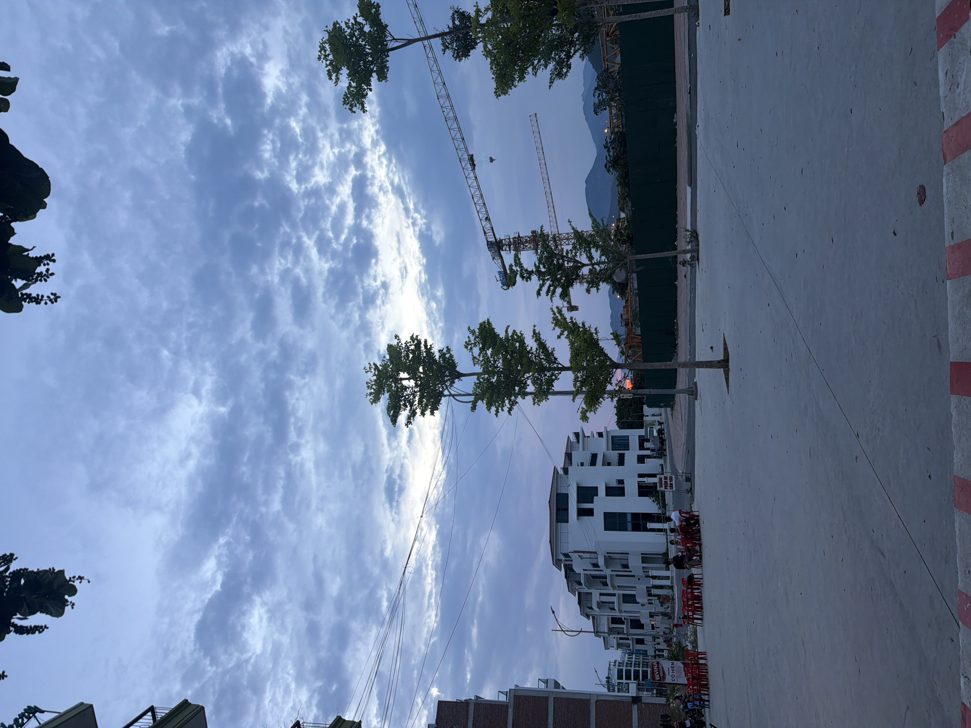 The image captures an exterior scene in Nha Trang Ward, Vietnam, during what appears to be dusk or early morning. The sky is filled with dramatic, cloudy formations, colored with hues of blue and gray, with hints of brighter light suggesting the sun is low on the horizon. In the mid-ground, a row of modern white buildings with dark-framed windows is visible, suggesting residential or commercial properties. To the left, a brick building with balconies is partially visible. In front of the white buildings, several red plastic chairs and tables are set up, with a few individuals seated, engaged in conversation. The foreground is a paved open area, possibly a plaza or street, with a red and white striped border. In the background, construction cranes dominate the skyline, indicating ongoing development. A green fence runs along the construction site, with a distant view of mountains. The overall impression is one of a developing urban area transitioning from day to night, with a blend of completed structures and ongoing construction.