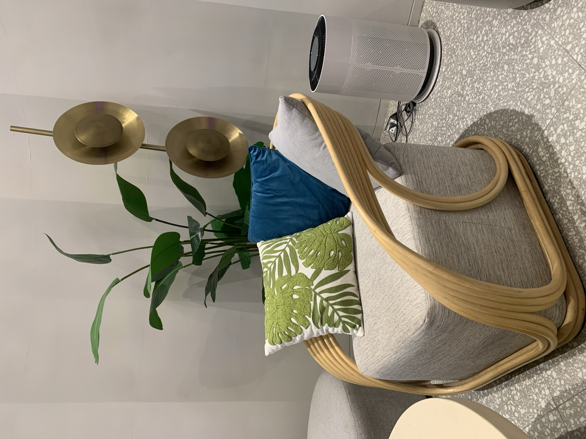 This is a close-up, indoor shot of a modern living space. The primary subjects are a grey upholstered armchair with a distinctive rounded rattan frame, and several decorative pillows. A large, floor-standing air purifier with a cylindrical white perforated body and a black top is positioned behind the armchair. To the left of the armchair, a brass wall sculpture with two circular discs is mounted on a white wall, and a tall potted plant with large, dark green leaves is situated in front of it. The armchair is adorned with three pillows: a light grey one, a deep blue velvet one, and a white pillow featuring an embroidered green monstera leaf pattern. The floor is a speckled terrazzo tile in shades of grey and white. The lighting appears to be artificial, creating soft shadows. The scene conveys a sense of contemporary, minimalist interior design with a touch of organic elements from the plant and rattan. There is no visible text in the image.