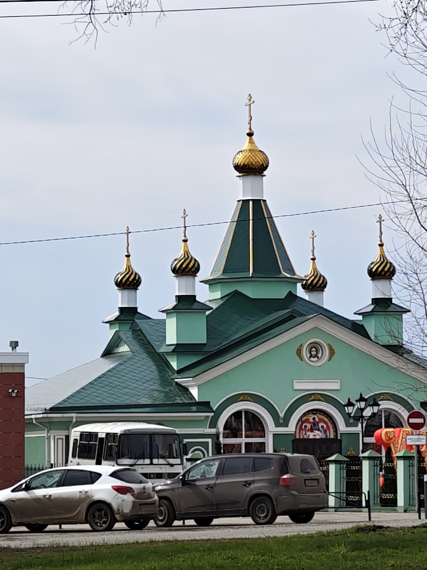 The image depicts a green and white Eastern Orthodox church in the city of Chaikovsky, Russia. The church features several golden domes with crosses atop them. A white bus, a dirty white car, and a dark grey SUV are parked in front of the church. A red building section is partially visible on the left. A no-entry sign is visible next to a gate with lanterns on the right of the image. Tree branches are seen against a pale sky.