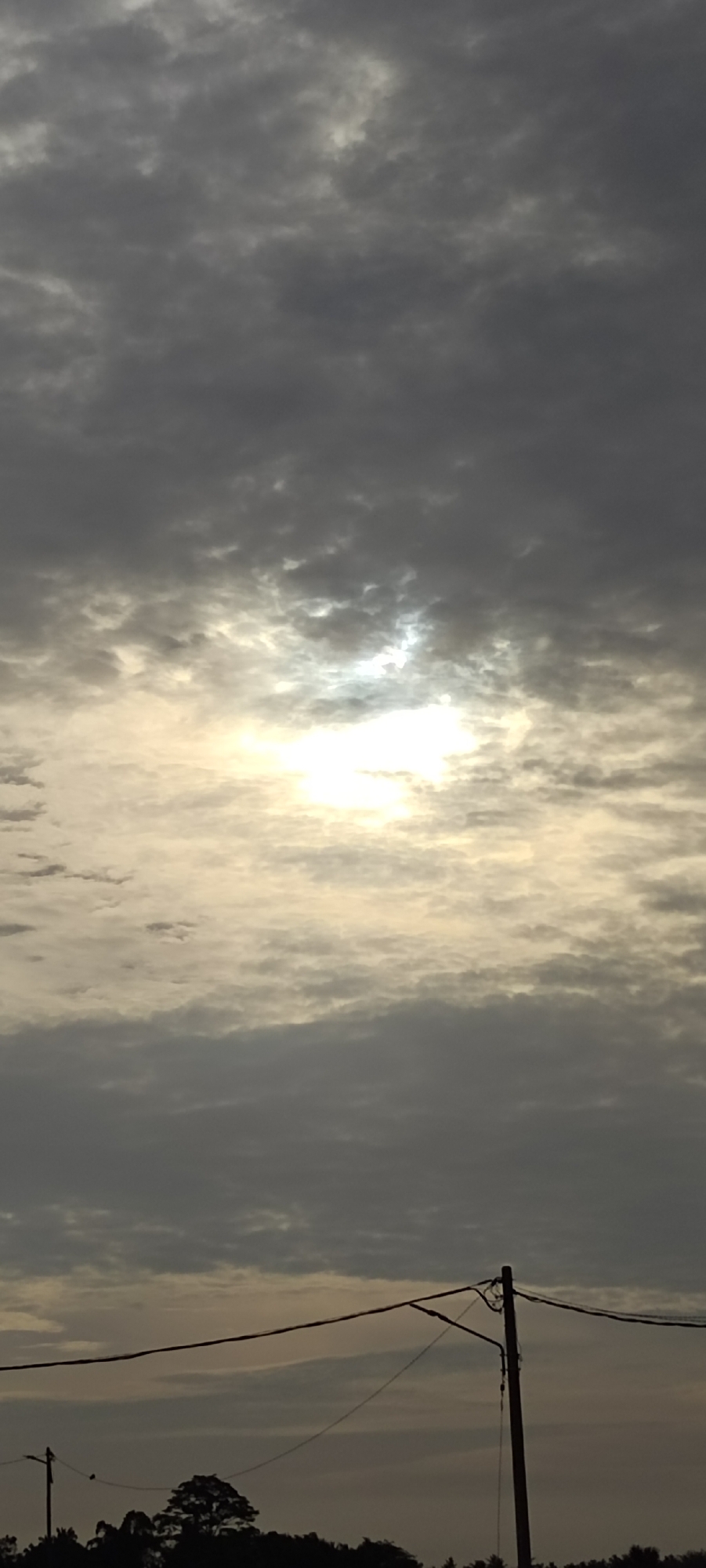 The image depicts a cloudy sky with the sun partially obscured, creating a diffuse light effect. Below the horizon line, a row of trees and utility poles with wires are silhouetted against the sky. The location is Meunasahaluedrienlhoksukon Barat, Indonesia. No people are visible.