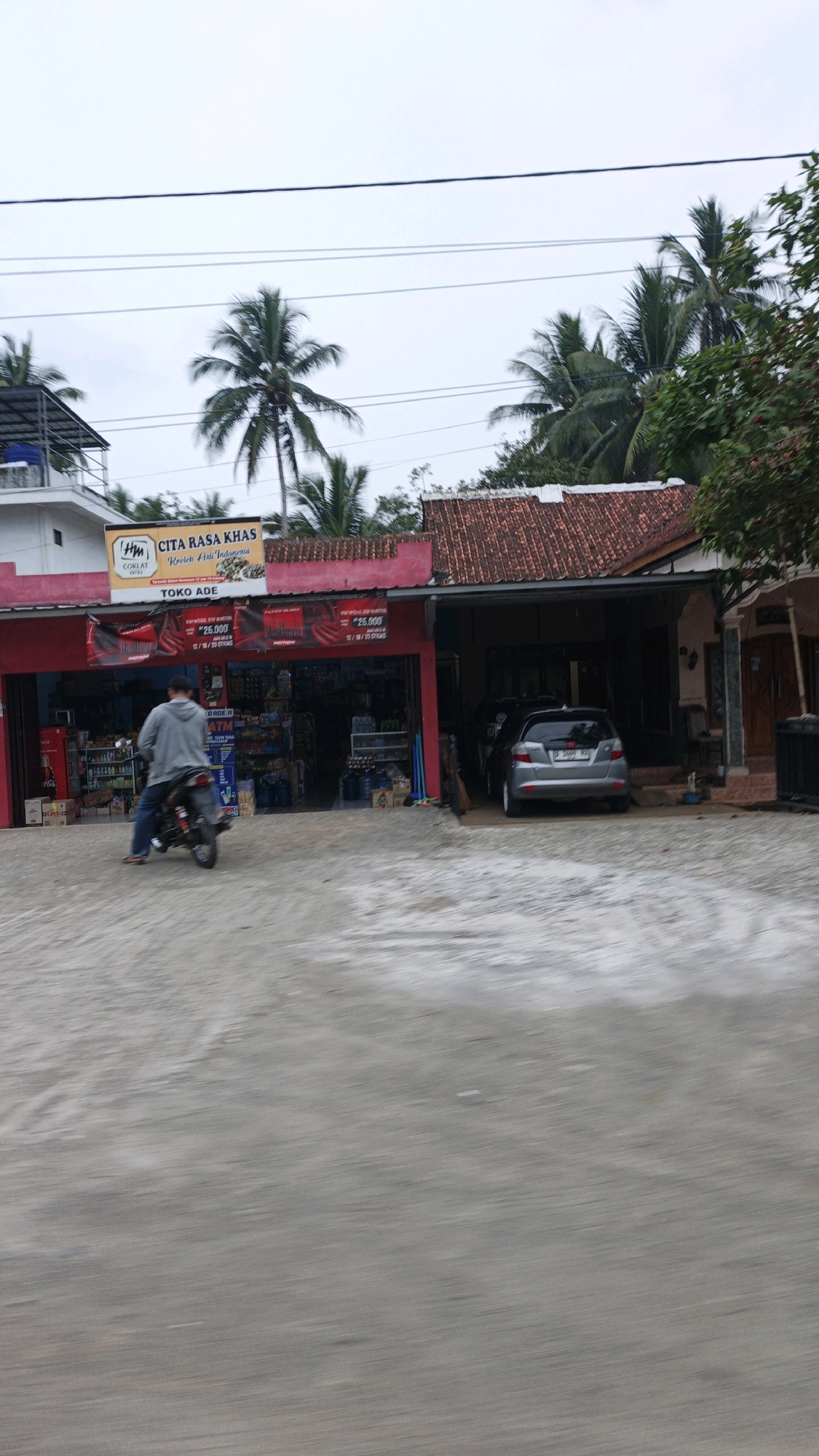 This is a medium shot, eye-level, outdoor, horizontal photograph taken in Cibenda, Indonesia, capturing a roadside scene. In the foreground, a light gray paved area with some white patches and debris is visible. A person is riding a motorcycle from left to right, wearing a light gray hooded jacket and blue jeans. The motorcycle is dark colored with a visible license plate and is moving.
The middle ground features a commercial establishment with a red facade and a sign that reads "CITA RASA KHAS" (Special Taste) with a logo that includes "COKLAT" (Chocolate) and the text "Kueleh Adli Indonesia" (Adli's Indonesian Cake). Below this sign is another sign that says "TOKO ADE" (Ade's Shop). The storefront appears to be a retail shop with shelves stocked with various items visible through the open front. There is a red refrigerator on the left side of the entrance and stacks of bottled water on the right. A silver car is parked in front of an adjacent building to the right, which has a tiled roof and a wooden door. The car has a license plate that reads "B 2099 XA".
The background consists of several palm trees and other greenery, suggesting a tropical or subtropical environment. The sky is overcast and gray, indicating a cloudy day. Electrical wires are strung across the top of the image. The overall scene depicts a typical day at a small roadside shop in an Indonesian town.