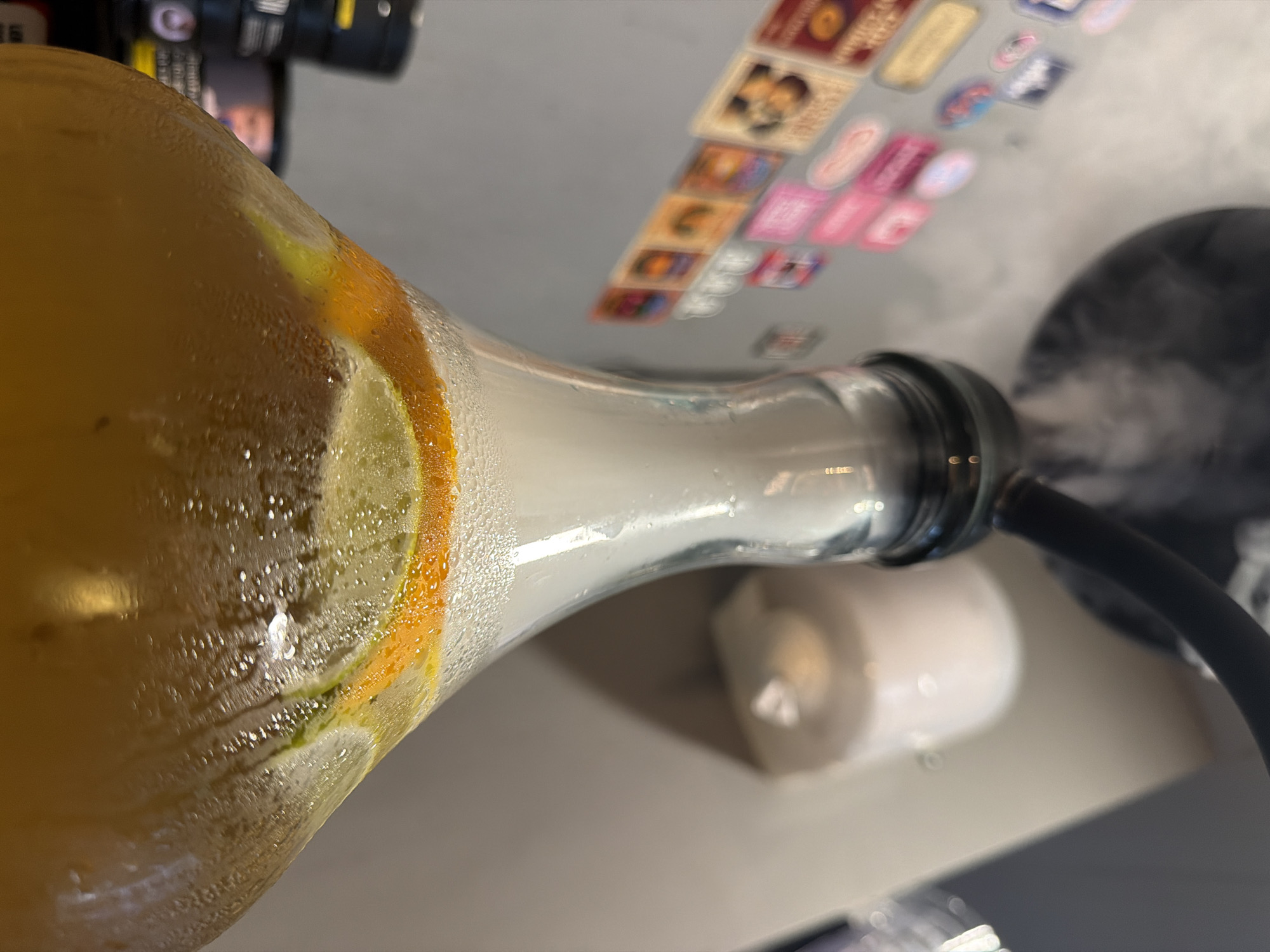 The image shows a close-up view of a partially filled glass bottle, likely a beverage container, with slices of citrus fruit inside (orange and perhaps lime or lemon). The bottle appears to have condensation on the outside. The neck of the bottle is connected to a black hose, possibly part of a hookah or shisha setup, with white smoke visible near the hose's opening. In the blurred background, there's a laptop covered with stickers, a white cylindrical object (possibly a candle), and what appears to be a camera lens. Given the specific and limited information, it is not possible to determine its exact location within Varna, Bulgaria.