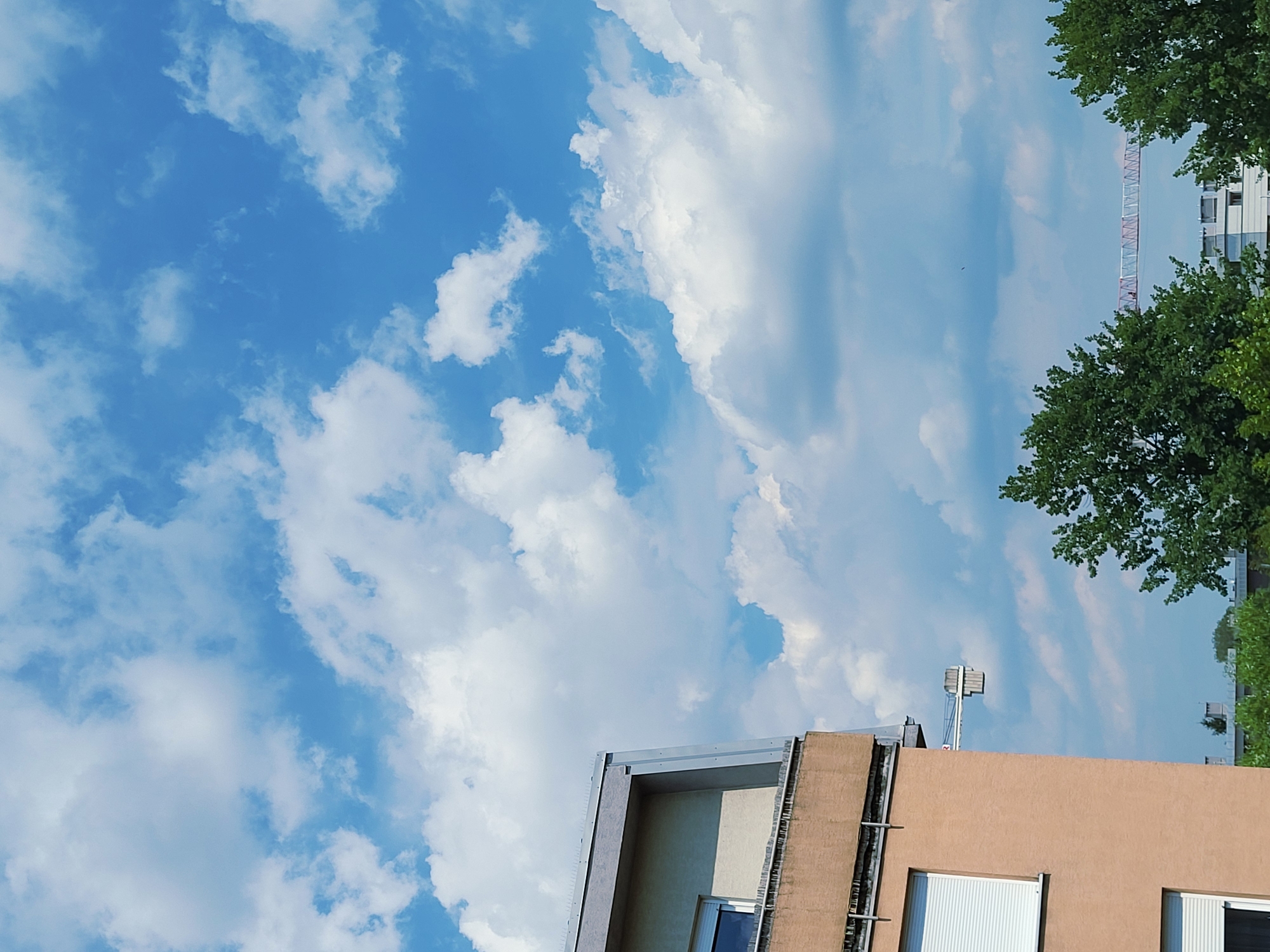The image shows a view of a city skyline with a building in the foreground and a bright blue sky with scattered white clouds above. The building is partially visible on the left side of the frame, with a peach-colored facade and a window with white blinds. A section of a crane, consisting of a boom and counterweight, is visible extending horizontally from the building. In the background, more buildings and trees can be seen, and a portion of a red and white construction crane stretches across the horizon. The scene appears to be captured on a sunny day.