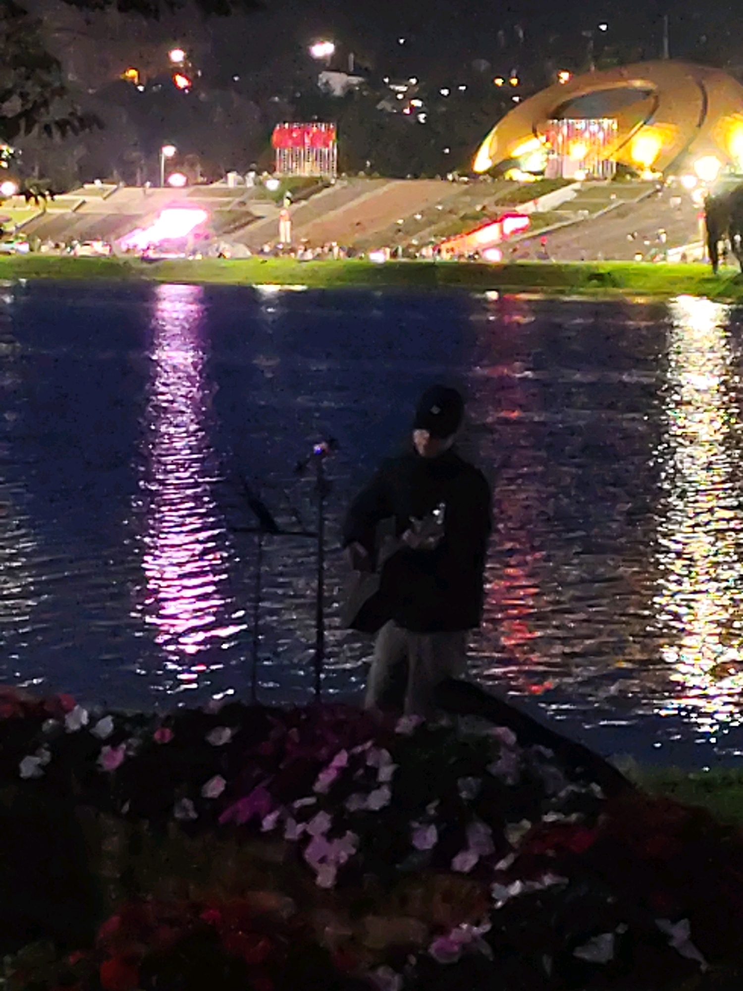 This image depicts an outdoor evening scene with a person playing a guitar by a body of water. The person is wearing a dark long-sleeved shirt, a baseball cap, and light-colored pants. They are standing on the edge of a grassy area, with a microphone stand positioned in front of them. The water reflects colorful lights from the background, which appears to be a city landscape with illuminated structures and buildings. The overall impression is of a performance or a relaxed evening gathering. The presence of a large, circular, golden structure in the background and tiered seating areas suggests this location might be an outdoor amphitheater or a prominent landmark in Da Lat, Vietnam, possibly near Xuan Huong Lake. The foreground features dark foliage and some scattered flowers, adding depth to the scene.